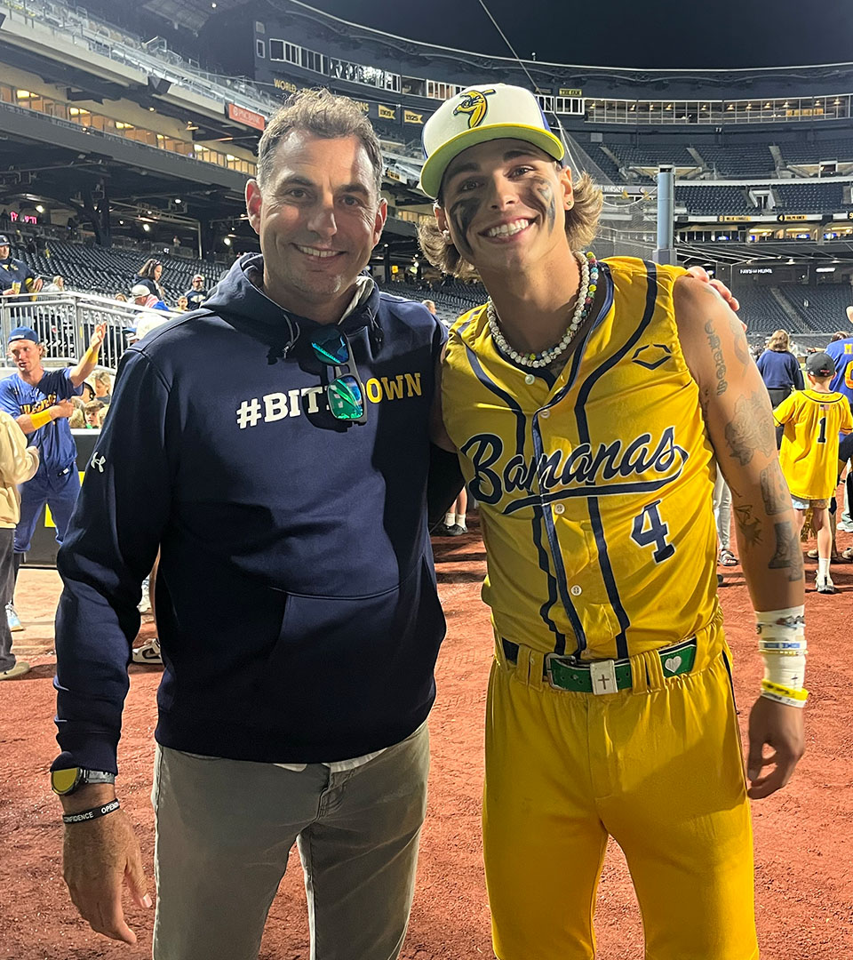 Kyle “KJ” Jackson of the Savannah Bananas stands smiling with Âé¶ąľ«Ńˇbaseball coach Jeff Duncan after a game, both posing on the field under stadium lights.