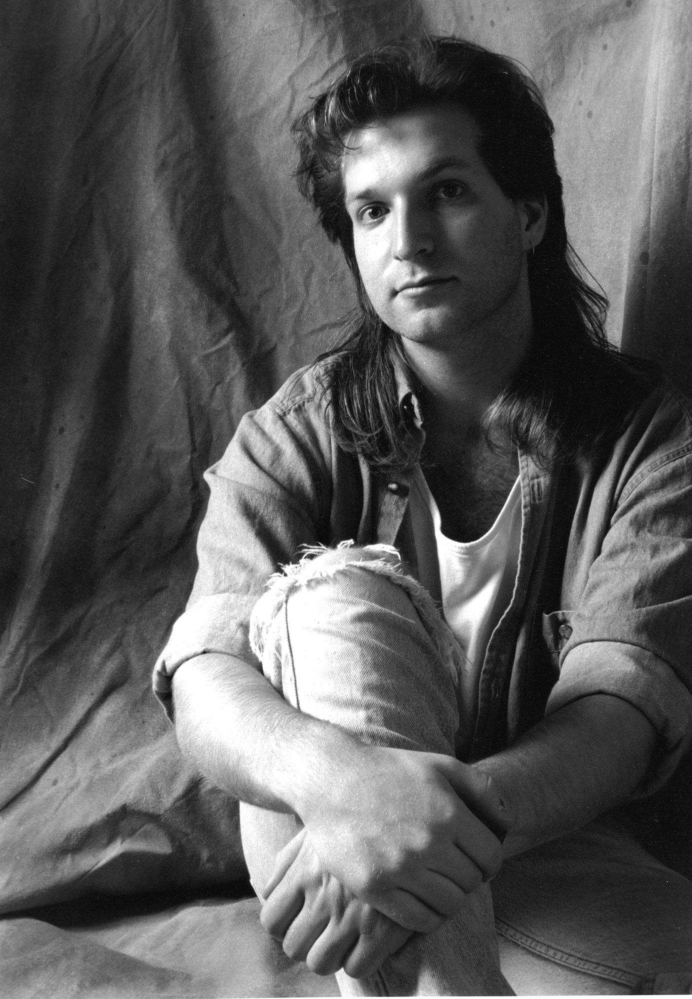 Black-and-white portrait of a young Marcus Lehto seated with his arms wrapped around one knee, looking slightly off camera.