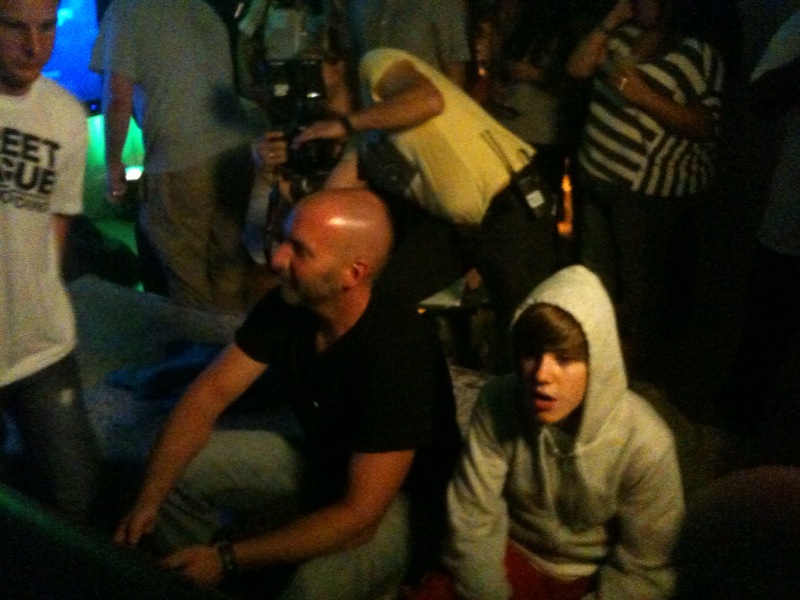 Marcus Lehto, seated and playing Halo Reach, next to Justin Bieber in a hoodie at a crowded event, with spectators gathered around them.