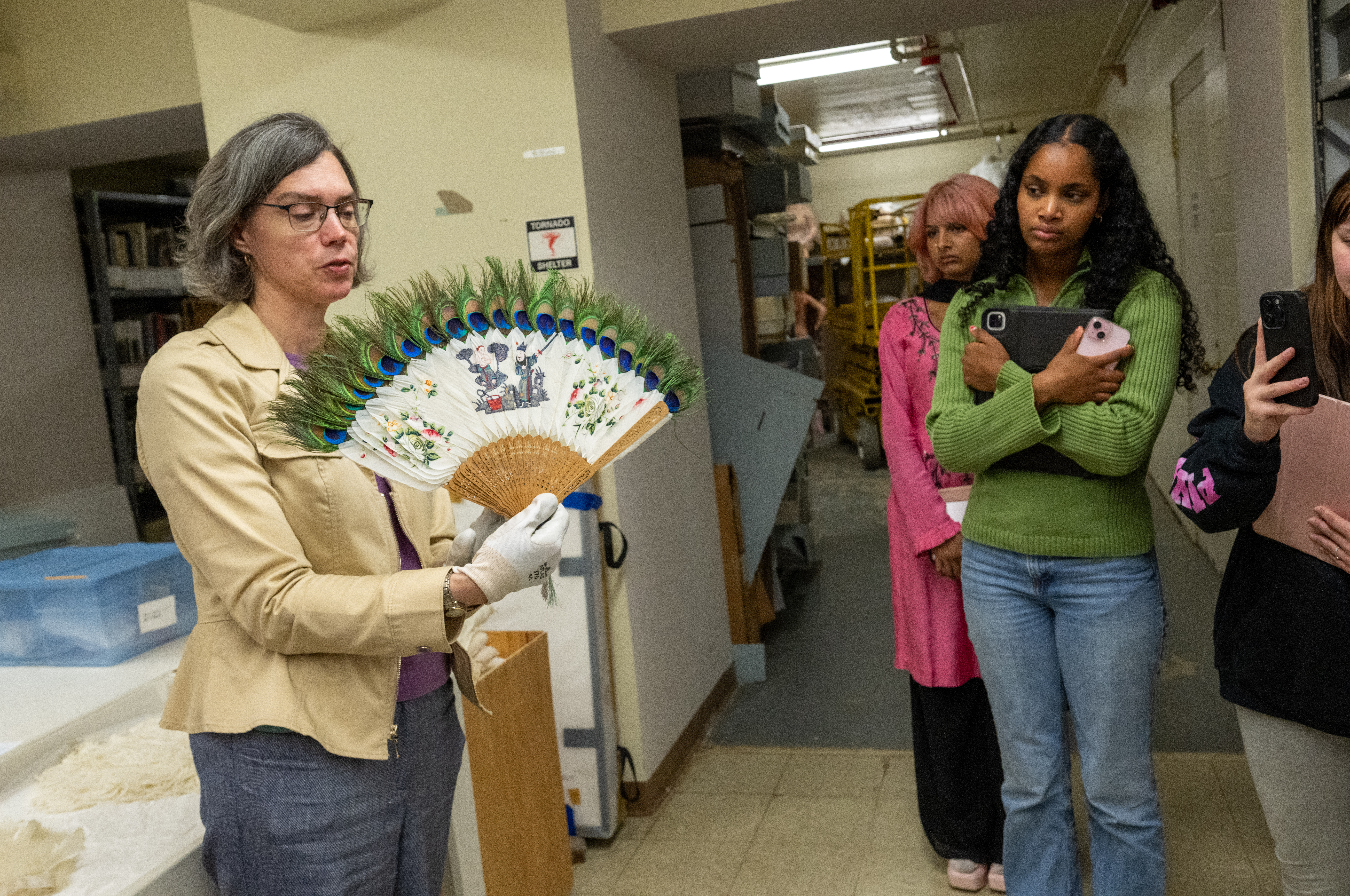 Woman with gray hair holds open a feathered fan while wearing white gloves in front of a group of students. 