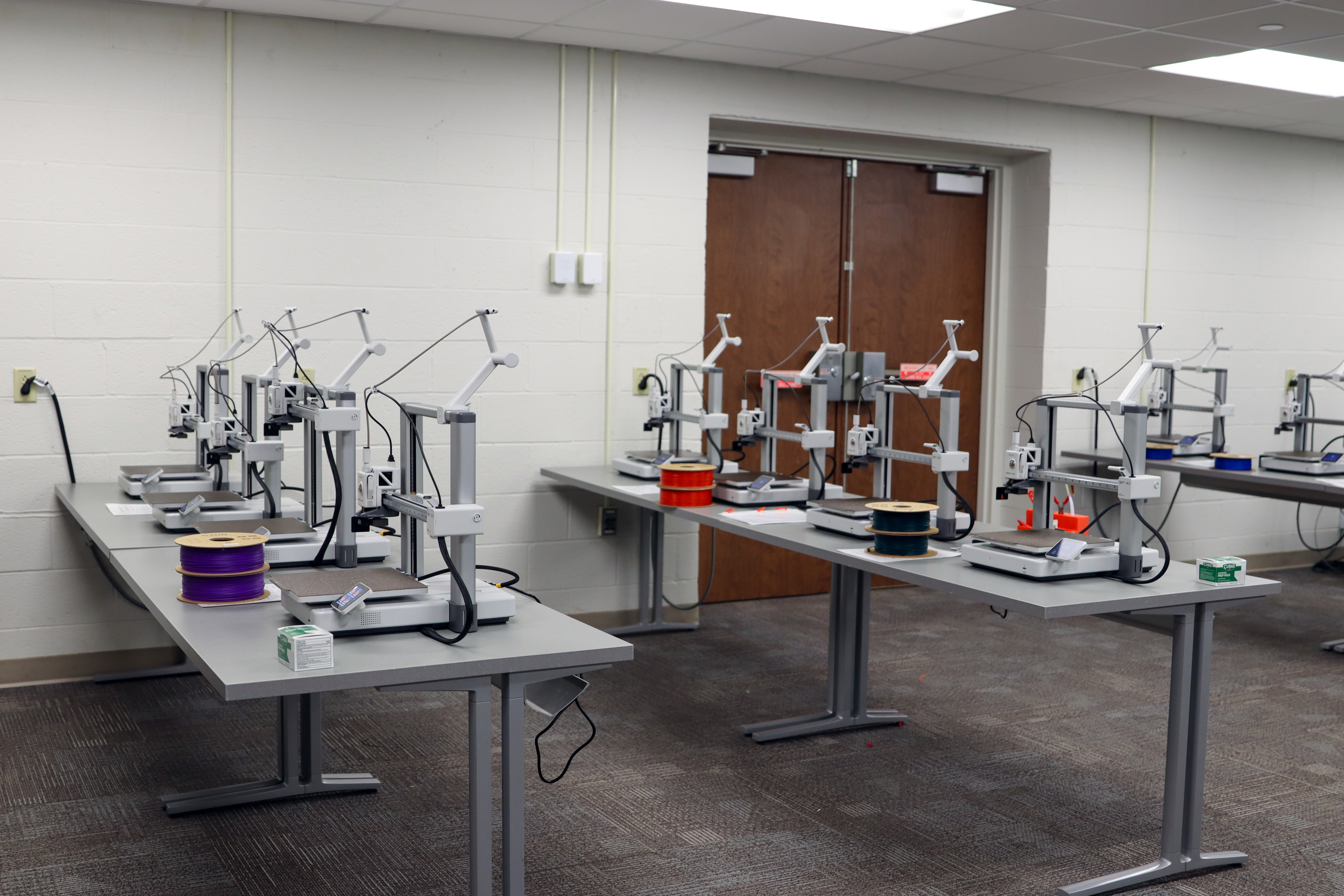 A classroom with multiple 3D printers arranged on gray tables in rows. Each printer has a spool of filament in different colors, such as purple, red, and green. The room has white walls, a carpeted floor, and double wooden doors in the back.