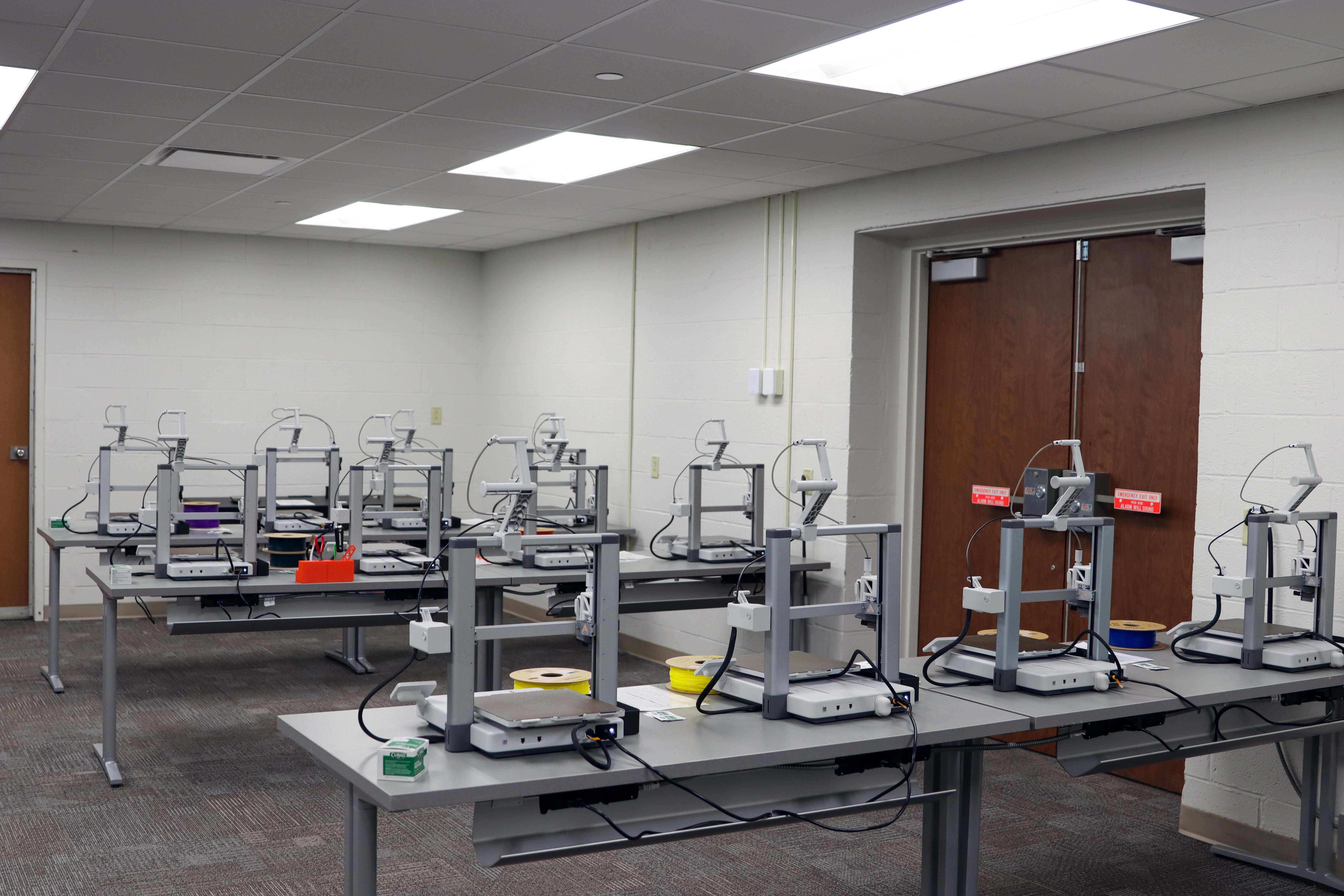 A classroom setup with multiple 3D printers arranged on gray tables in rows. Several printers have filament spools attached, including yellow, purple, blue, and green. The room has white walls, carpeted flooring, fluorescent ceiling lights, and double wooden doors at the back.