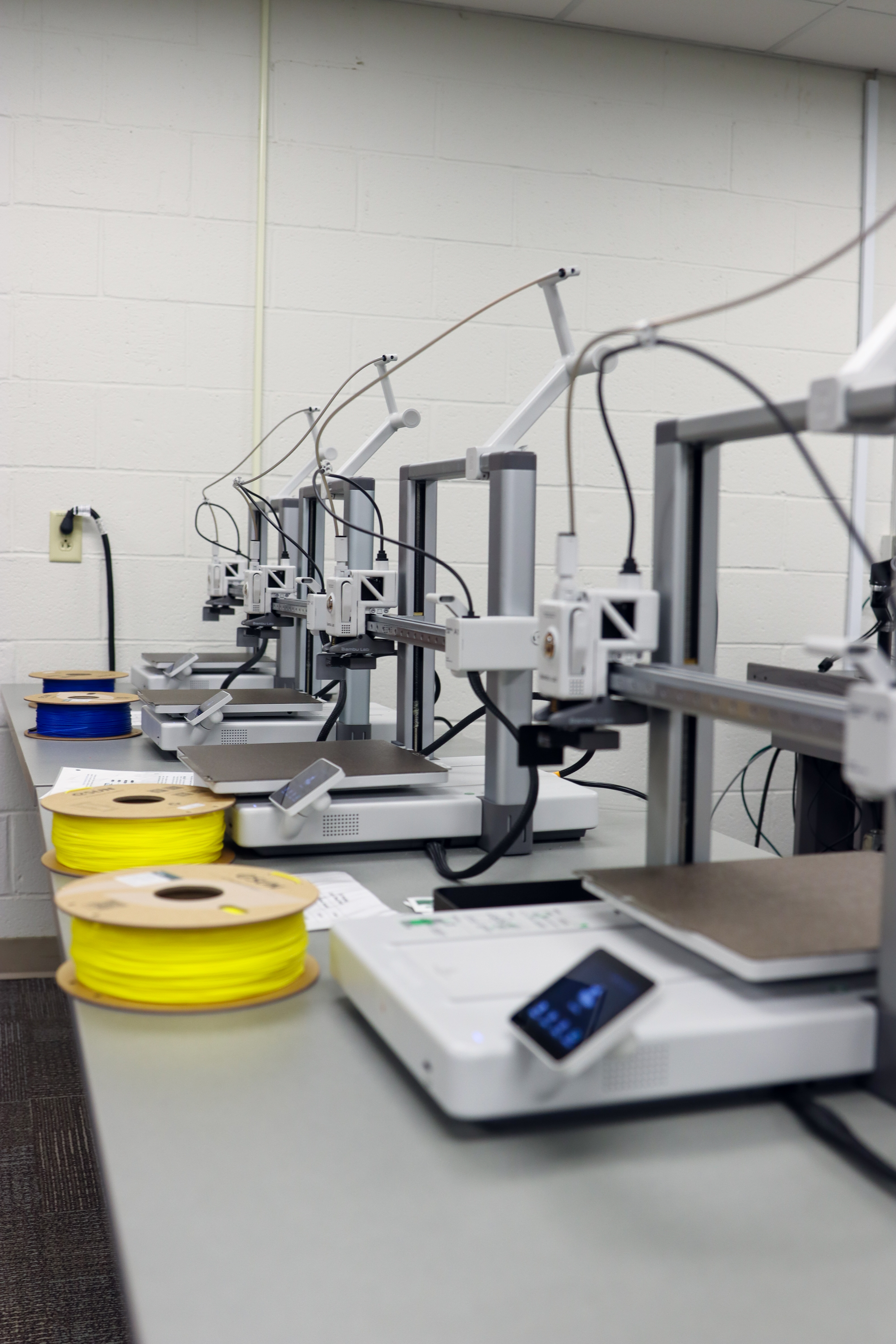 A row of desktop 3D printers lined up on a gray table in a classroom. Several spools of filament, including bright yellow and blue, are placed on the table near the machines. The printers have touchscreen controls and filament guides extending upward. The background shows a white cinder block wall and electrical outlets.