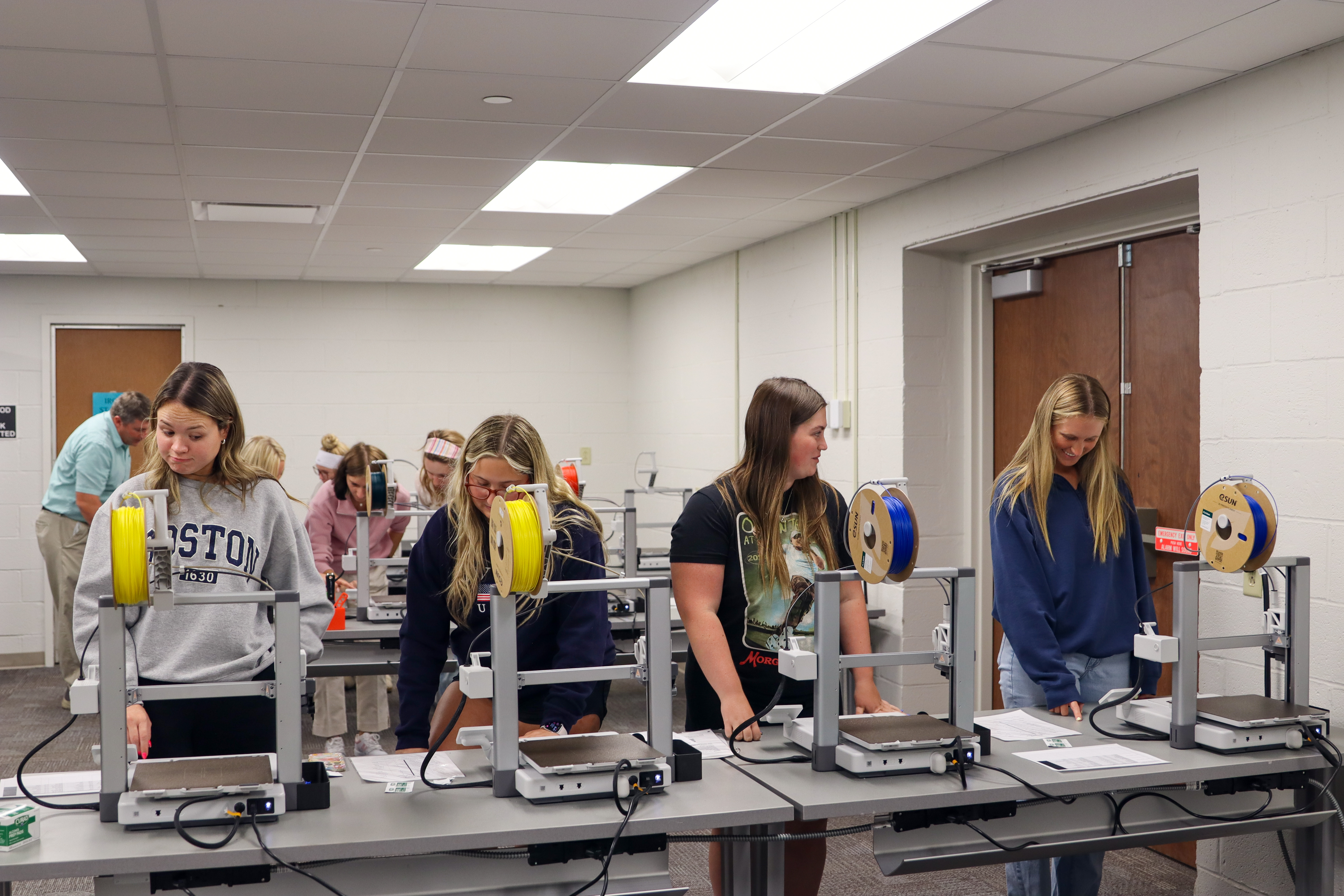 A group of students works in a classroom with several 3D printers lined up on tables. The printers have spools of yellow and blue filament loaded. Four students in the foreground are standing at the machines, reading instructions or adjusting settings, while more students and an instructor are visible in the background. The room has white walls, fluorescent lighting, and wooden double doors.