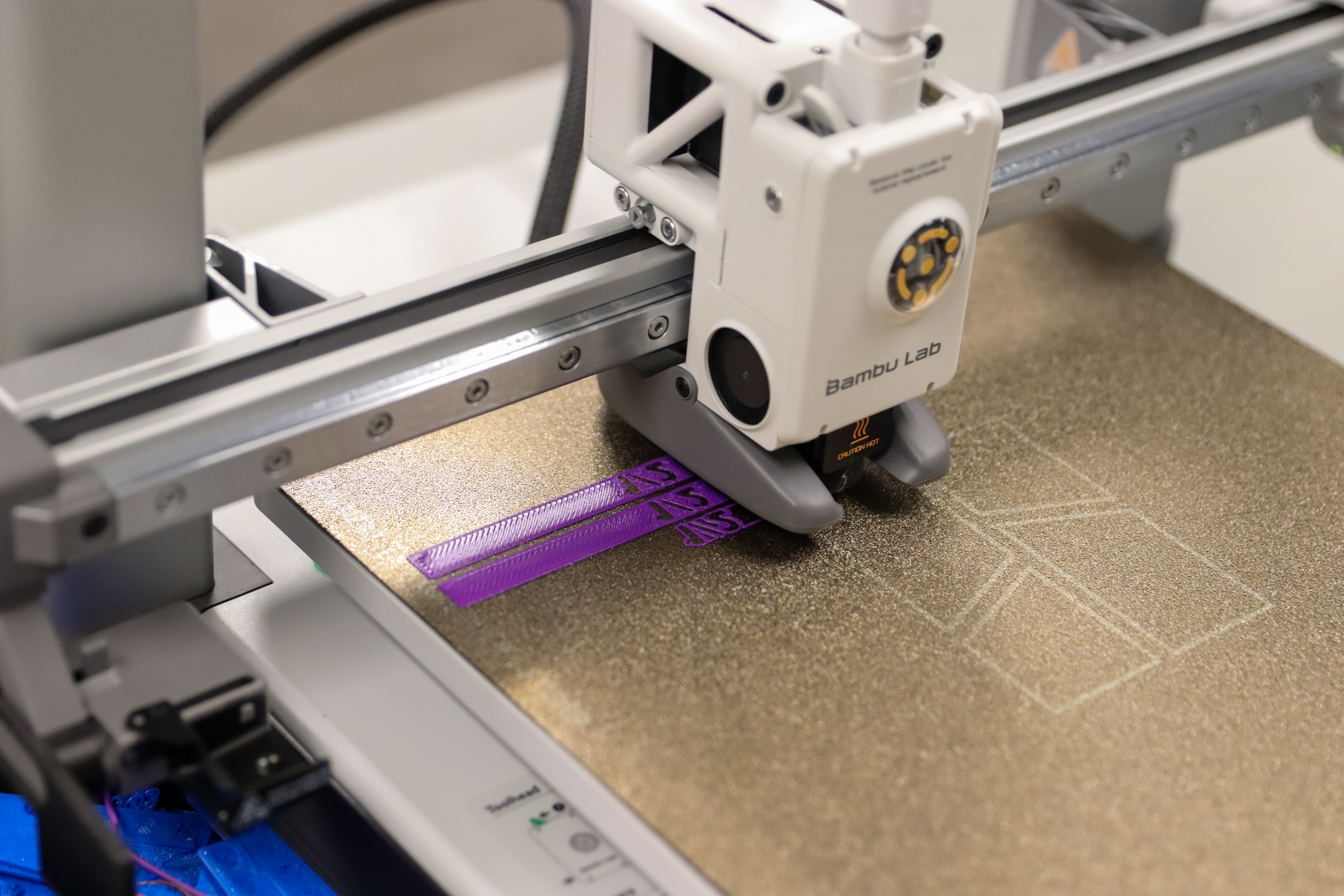 A close-up of a Bambu Lab 3D printer in operation, printing with purple filament. The printer nozzle is laying down layers on the build plate, forming two rectangular objects with visible infill patterns. The textured build surface also shows faint outlines of previous calibration prints.