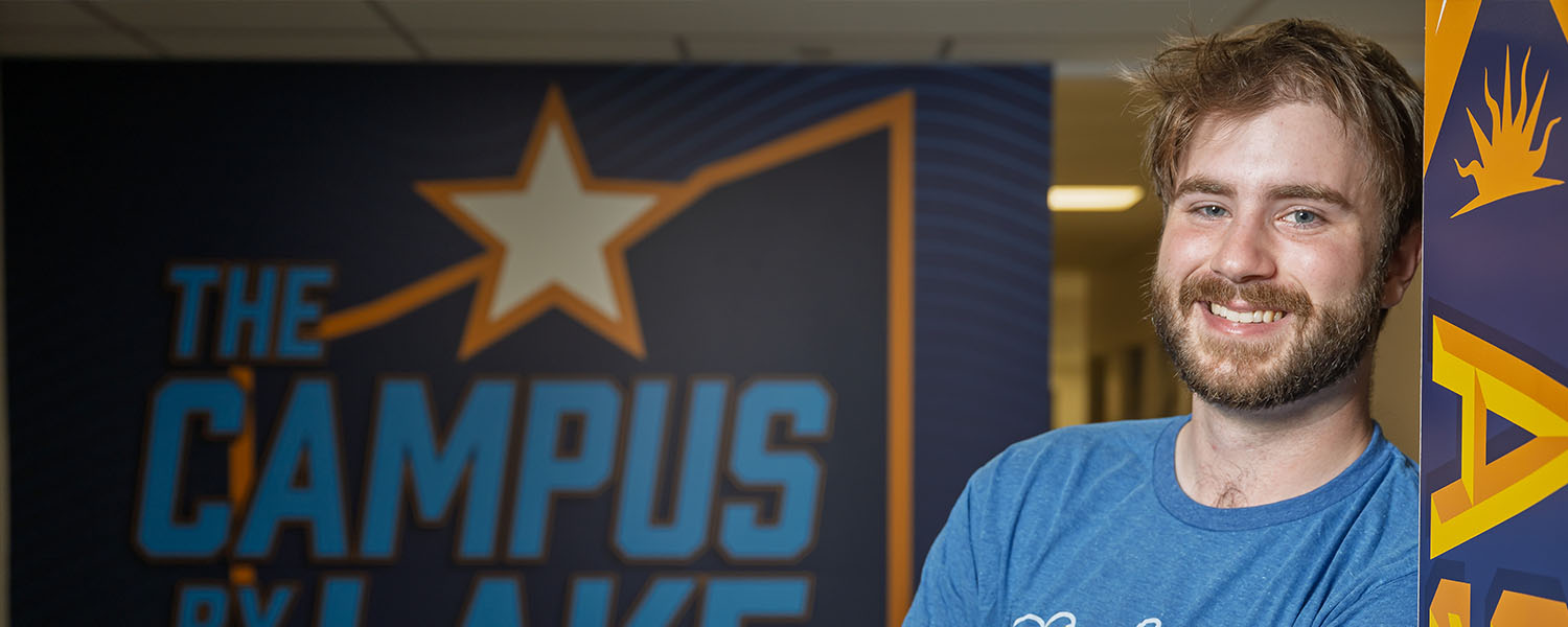 A smiling male student in a blue t-shirt leaning against a wall next to a large graphic that reads 'The Campus by the Lake' at Kent State University Ashtabula.