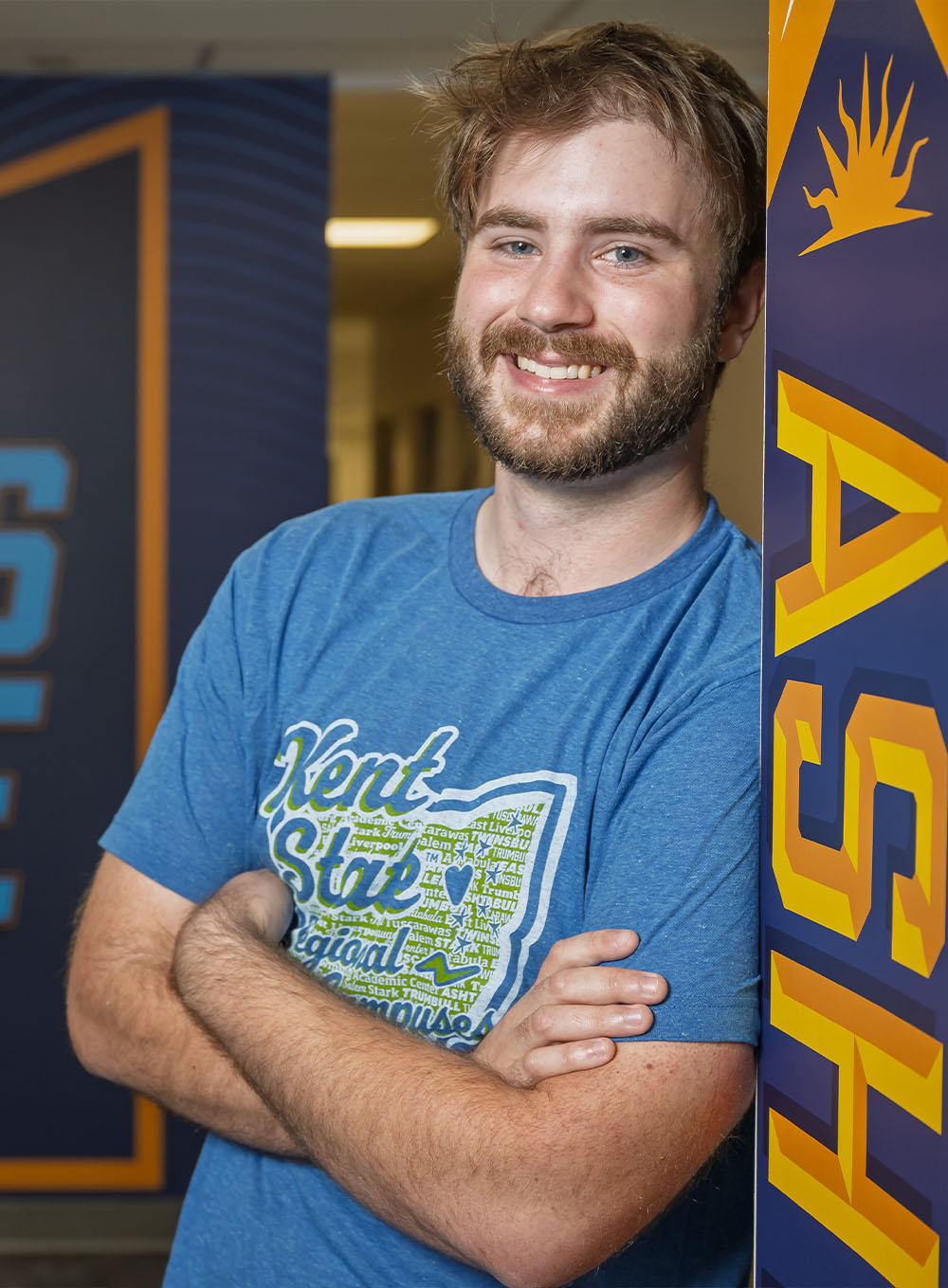 A smiling male student in a blue t-shirt leaning against a wall next to a large graphic that reads 'The Campus by the Lake' at Kent State University Ashtabula.