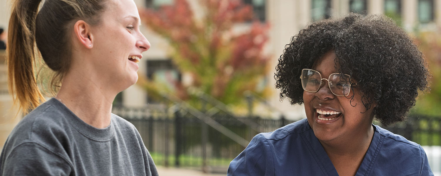 Four nursing students in blue scrubs sitting at a wooden outdoor table, talking and studying from textbooks at a Kent State Columbiana campus.