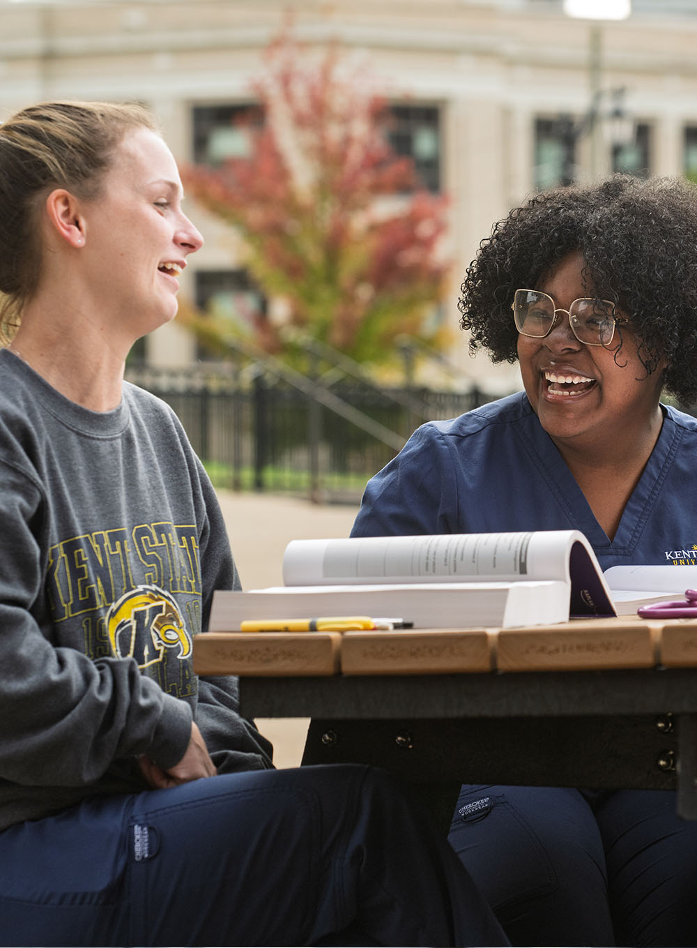 Four nursing students in blue scrubs sitting at a wooden outdoor table, talking and studying from textbooks at a Kent State Columbiana campus.