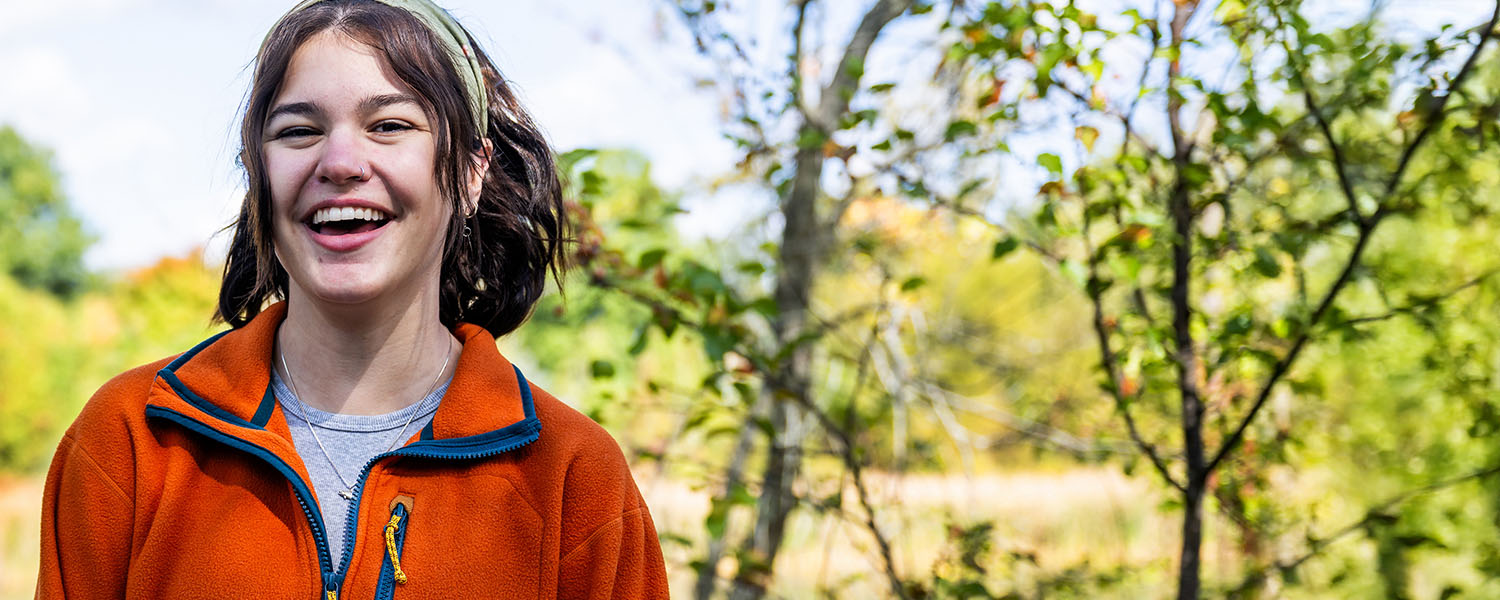 A female student in an orange fleece smiling outdoors in front of a pond and wooded area at Kent State University Stark.