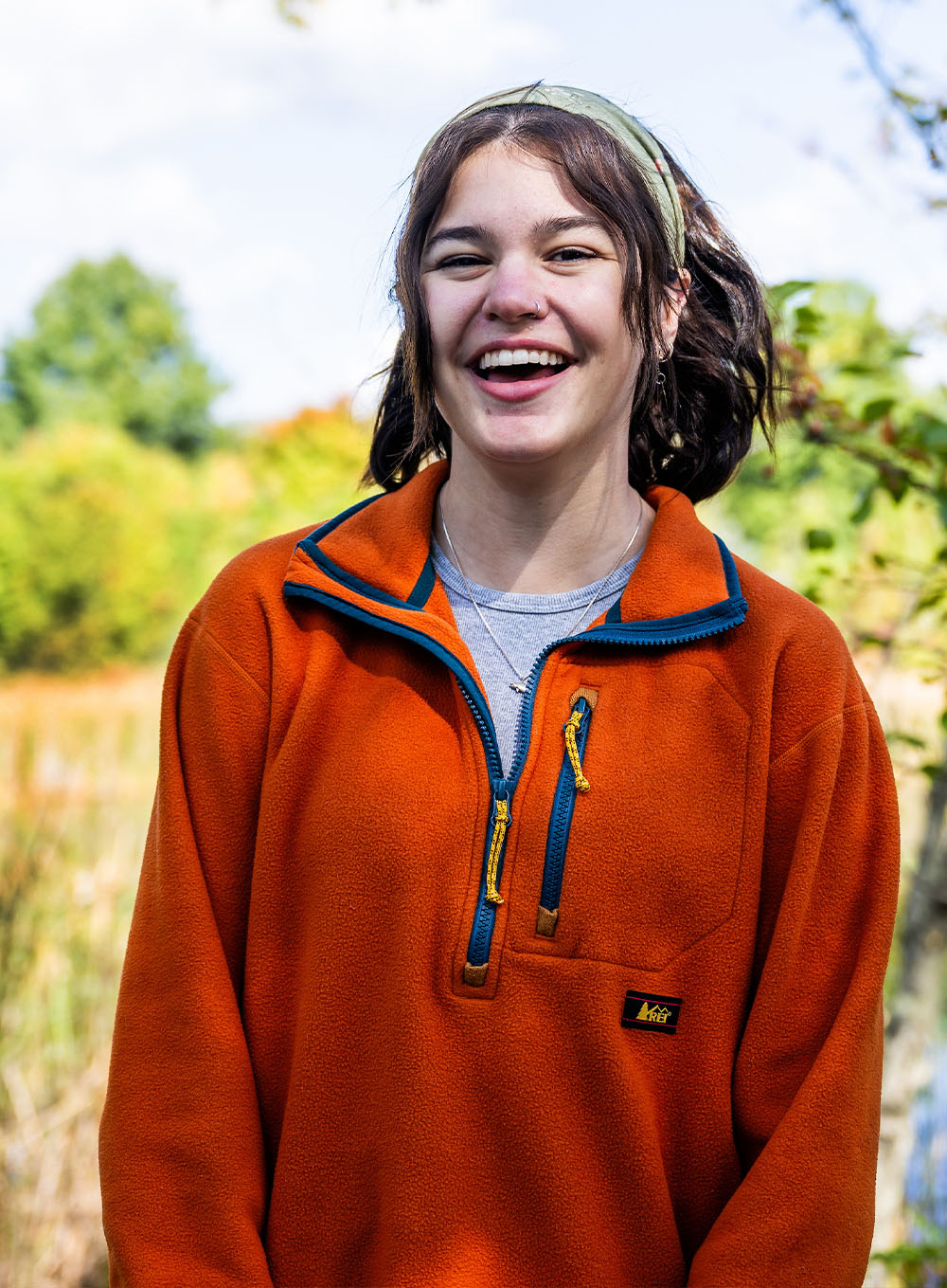 A female student in an orange fleece smiling outdoors in front of a pond and wooded area at Kent State University Stark.