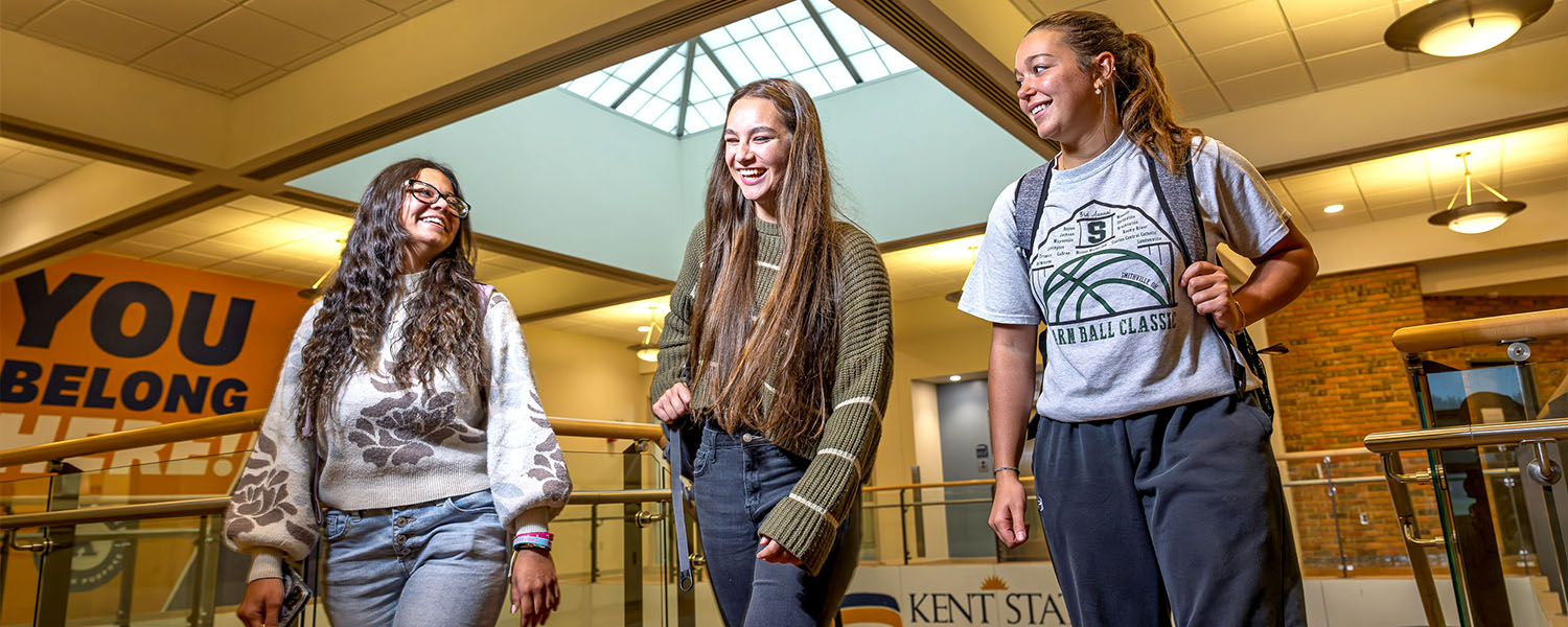 Three students walking and laughing on an indoor balcony at Kent State University Trumbull, featuring a 'You Belong Here' banner in the background.
