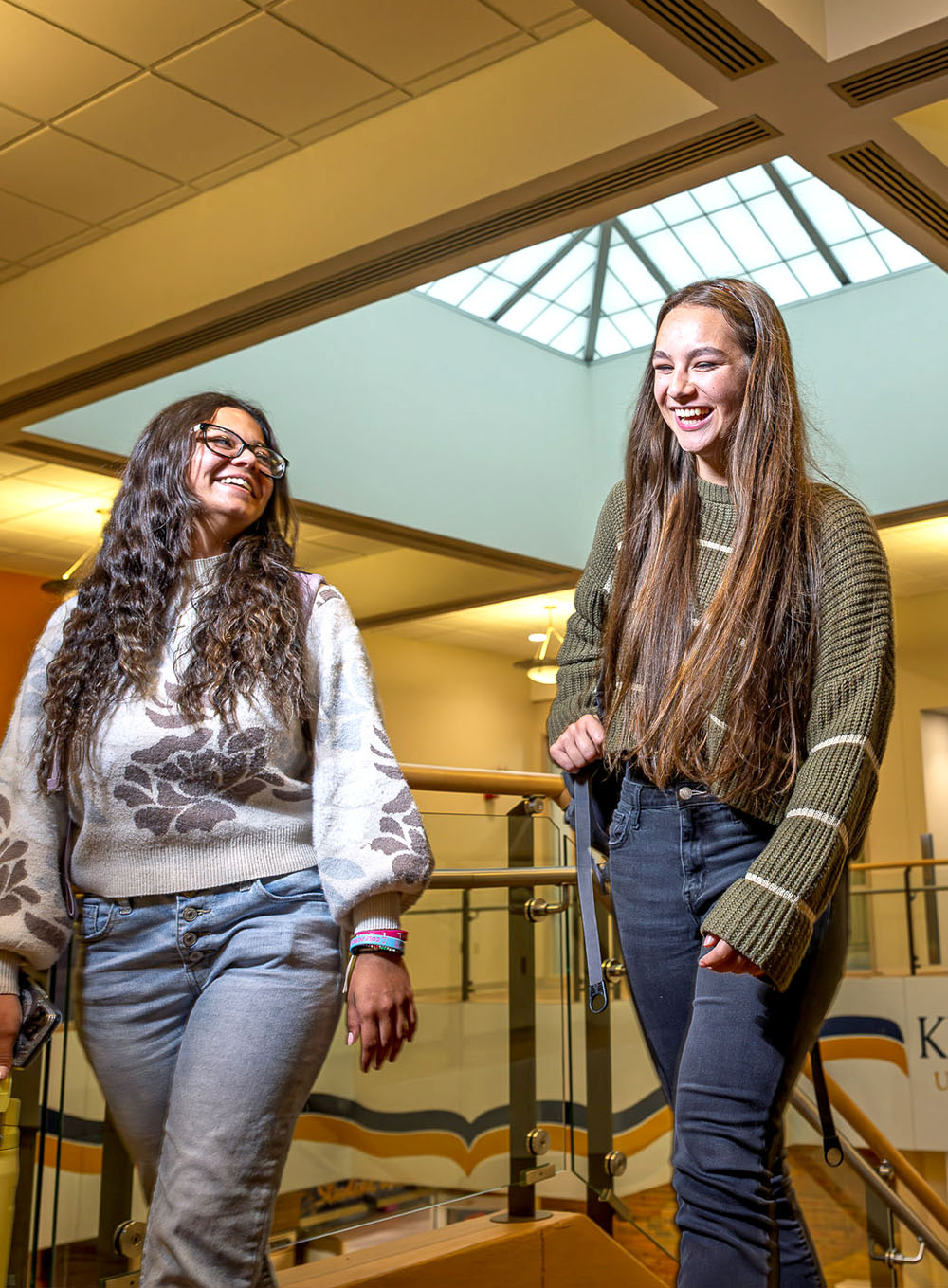 Three students walking and laughing on an indoor balcony at Kent State University Trumbull, featuring a 'You Belong Here' banner in the background.
