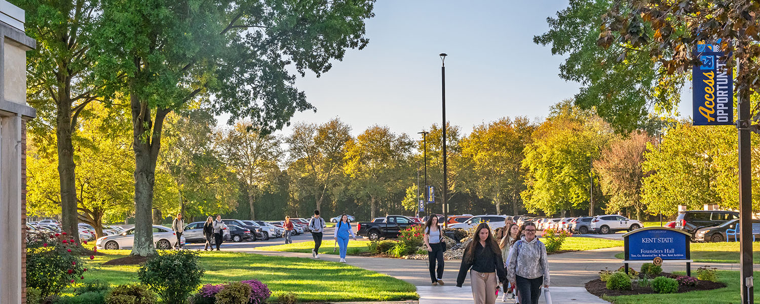 Students walking along a paved path toward Founders Hall at Kent State University Tuscarawas, surrounded by green lawns and autumn trees.