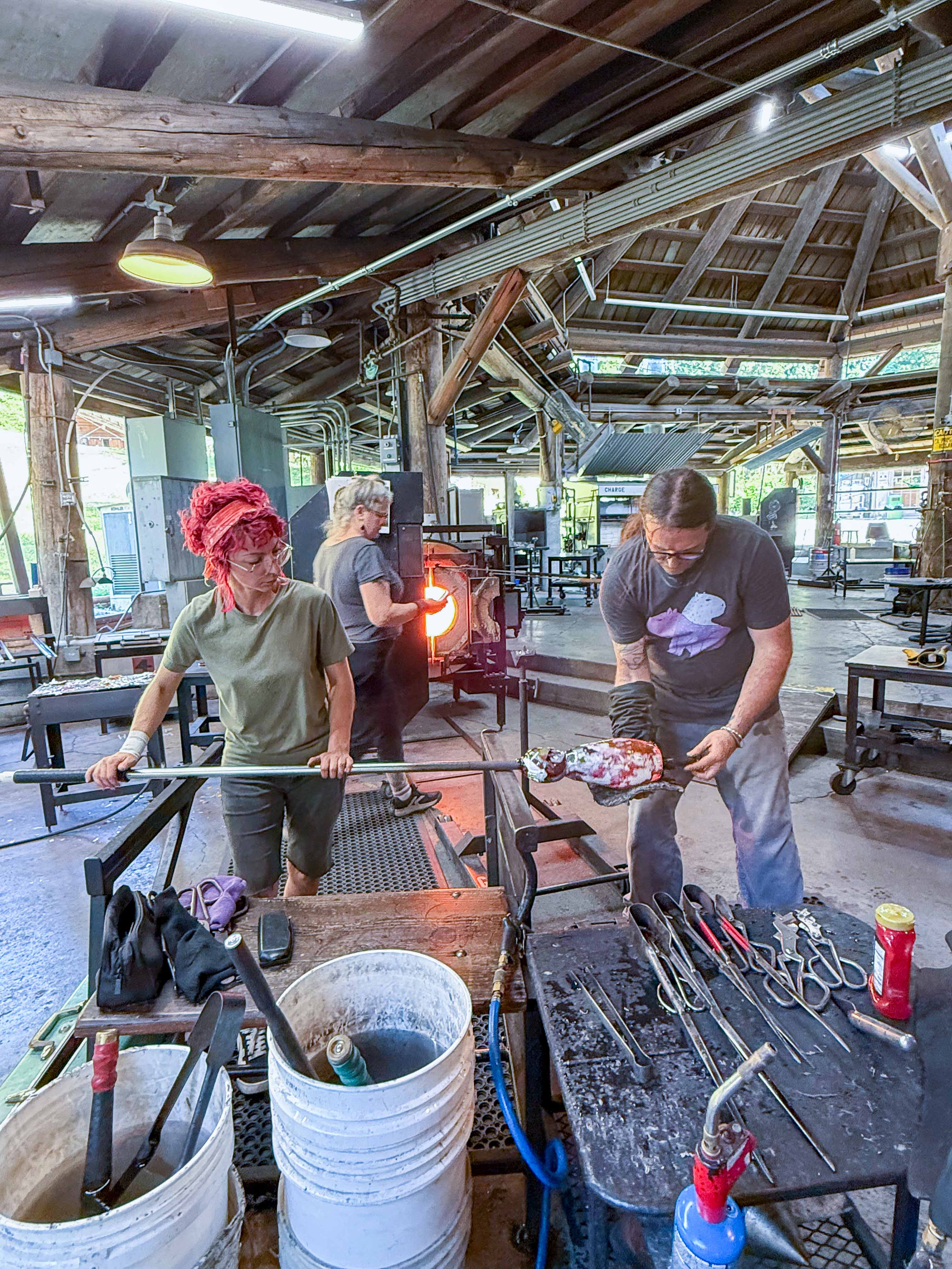 Wide view inside a glassblowing studio with a high wooden-beam ceiling and industrial equipment, showing multiple people working at separate stations; in the foreground, two people shape a glowing molten glass piece on a metal rod at a workbench covered with tools, while additional workstations and a furnace are visible in the background.