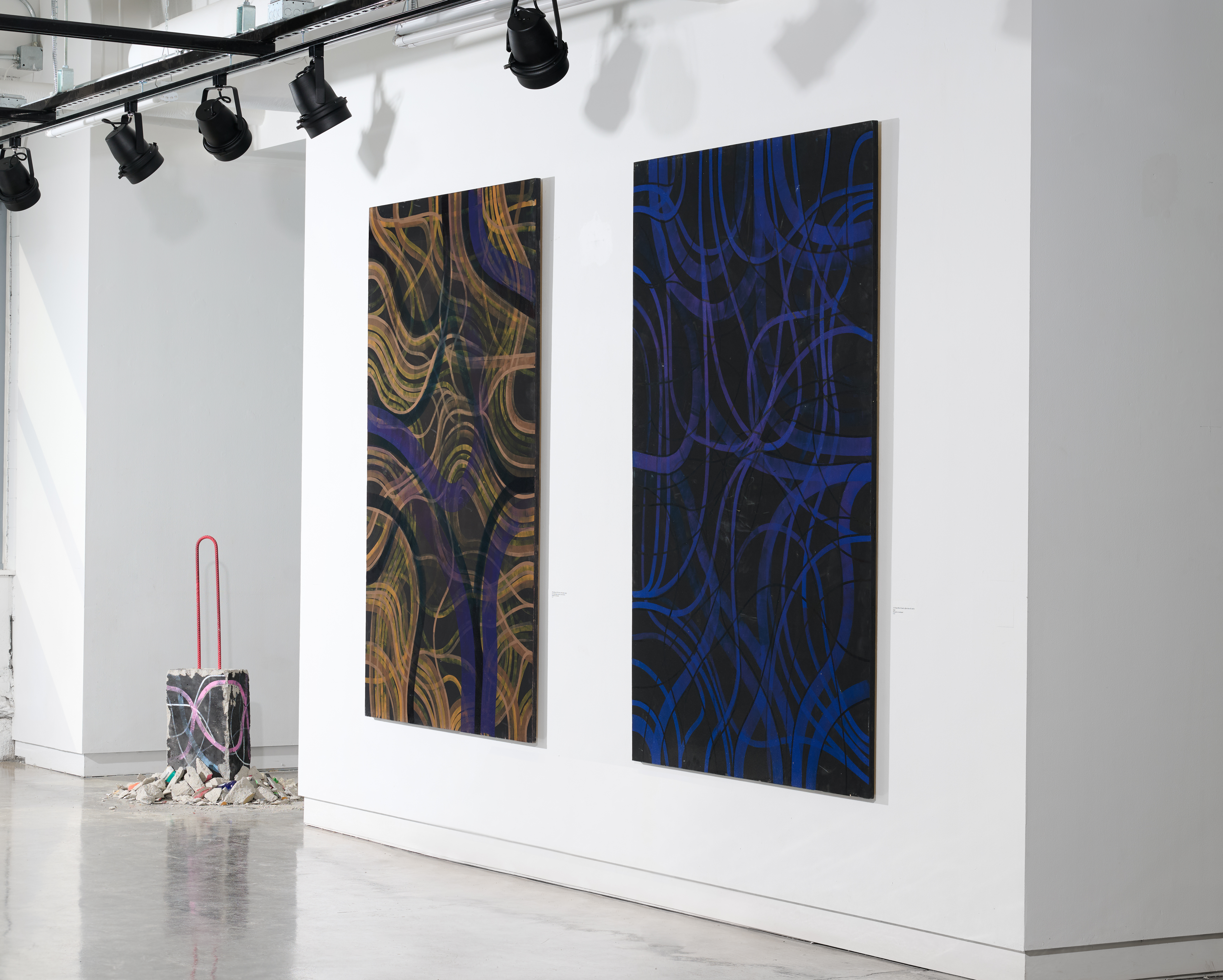 two multi-textured tapestrys on the wall of the gallery next to a painted chunk of cement on the floor surrounded by more crushed cement