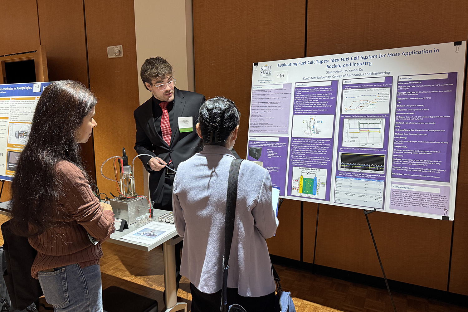 Two students listen to a presentation while standing in front of a large poster board.