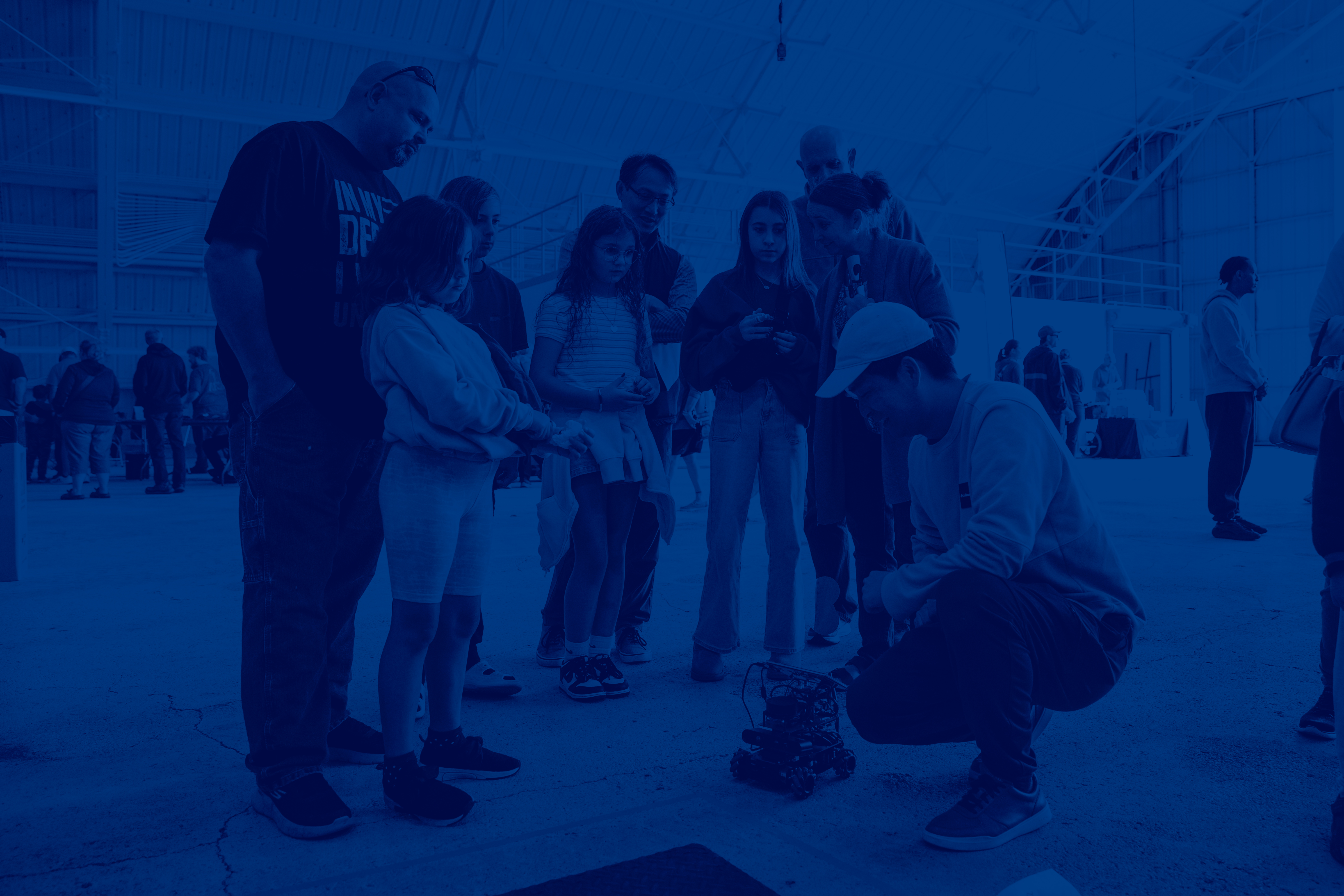 Children and parents watch an engineering student demonstrate a small wheeled robot.	