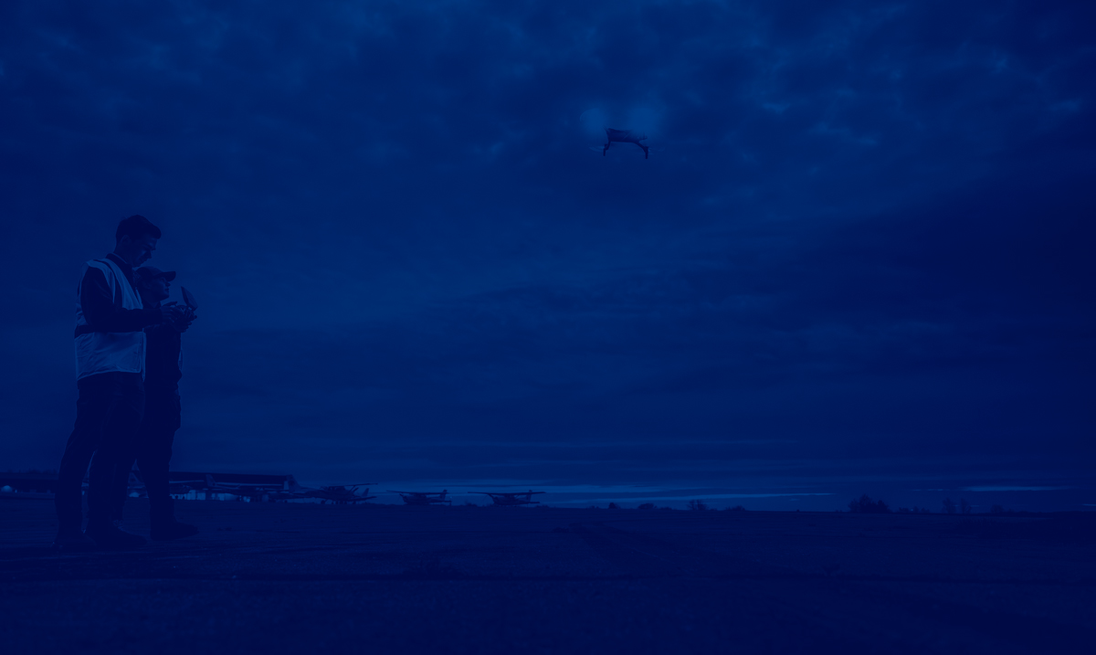 Two people stand outside piloting a glowing drone.	
