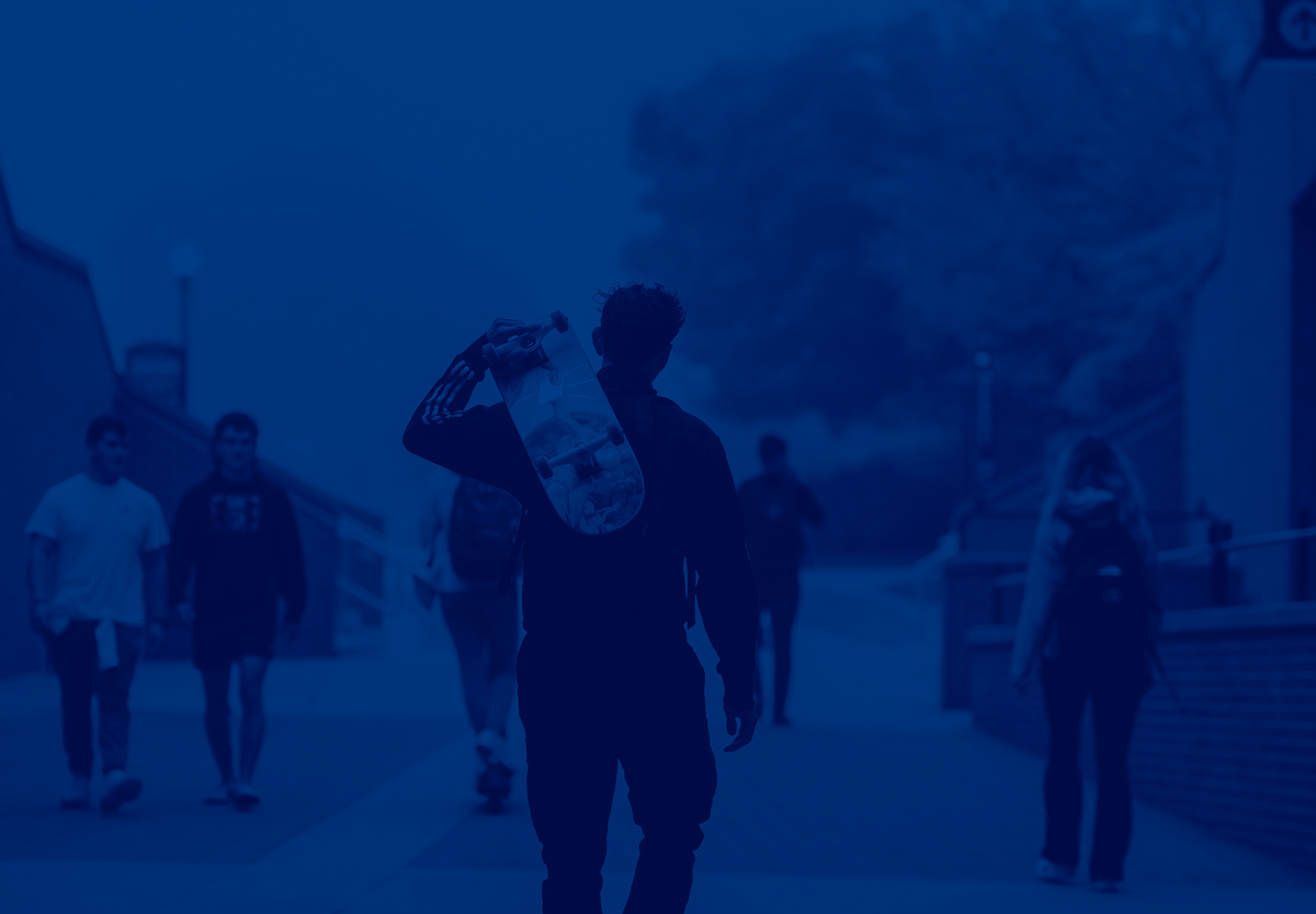 A student carrying their skateboard while walking on campus.	