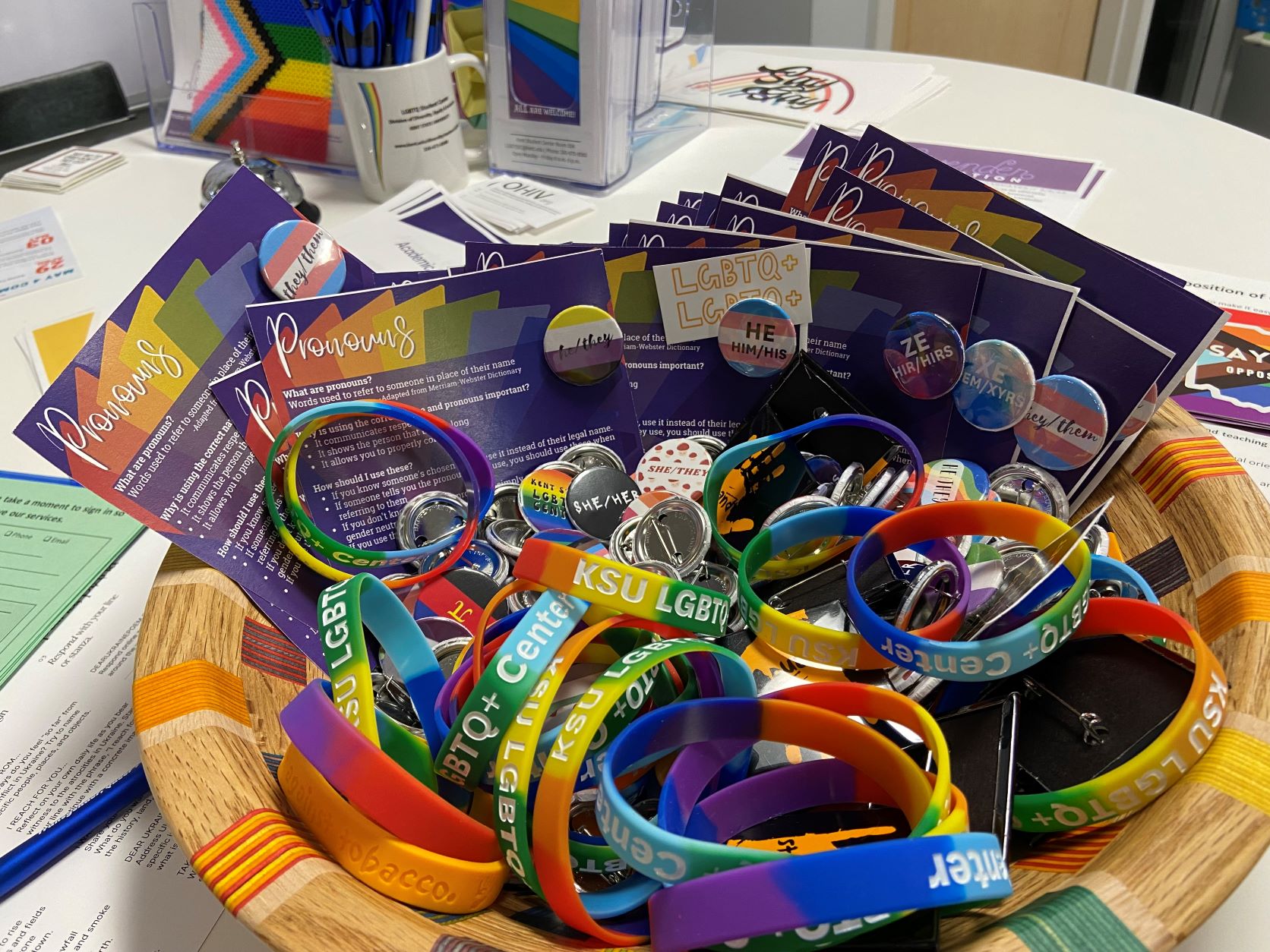 Picture of basket containing several pins of varying designs, all with different pronouns on them. There are also small informational sheets about pronouns, as well as rubber bracelets with words "LGBTQ+ Center" across the bands.