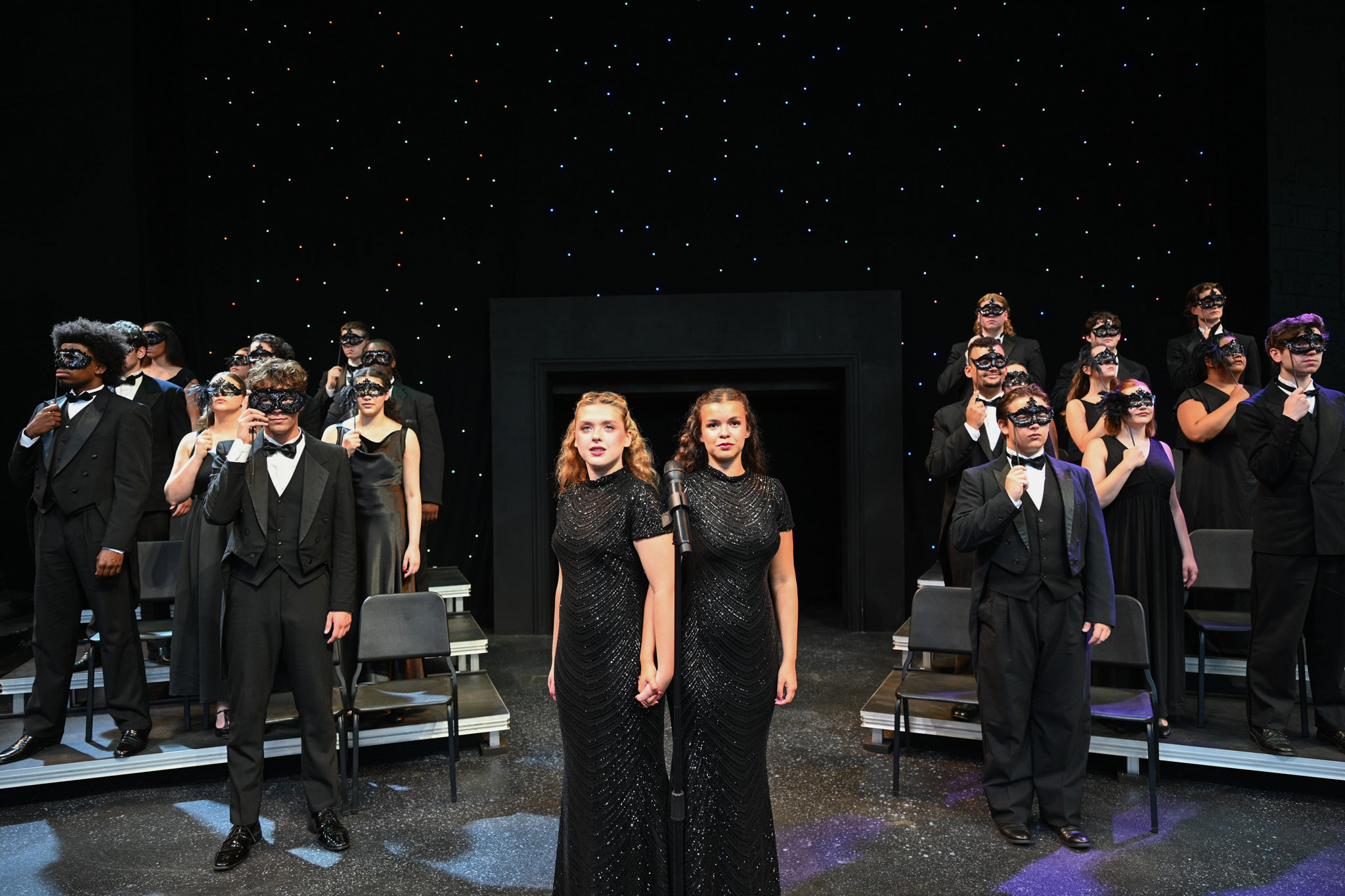 A cast and crew occupy a stage all dressed in black against a starry background. Taking center stage are characters Daisy and Violet, who wear long black dresses and stand conjoined at the hip