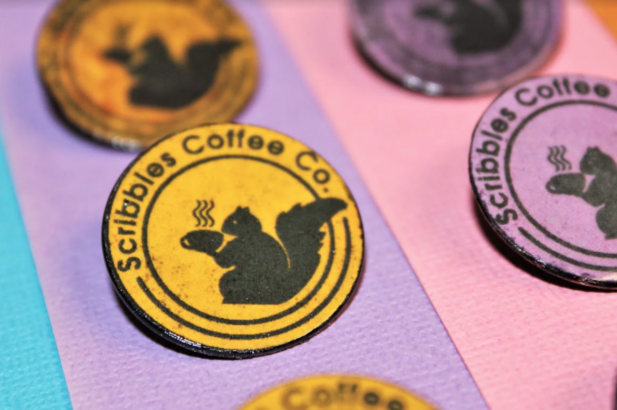 Round pins made by Danny Likar for Scribbles Coffee Co featuring a silhouette of a black squirrel drinking coffee.