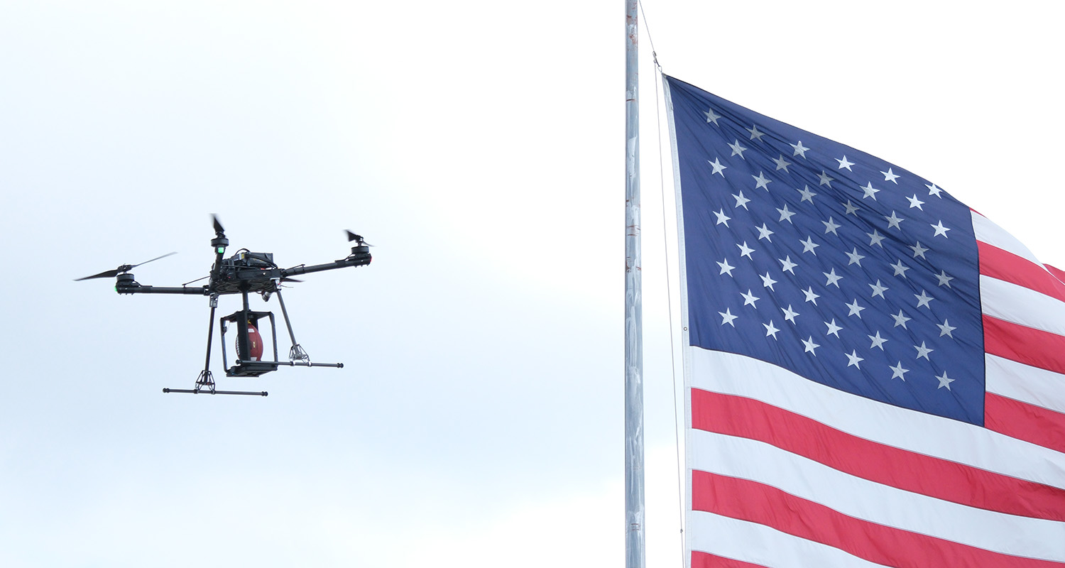 A quadcopter drone carrying a football in front of an American flag