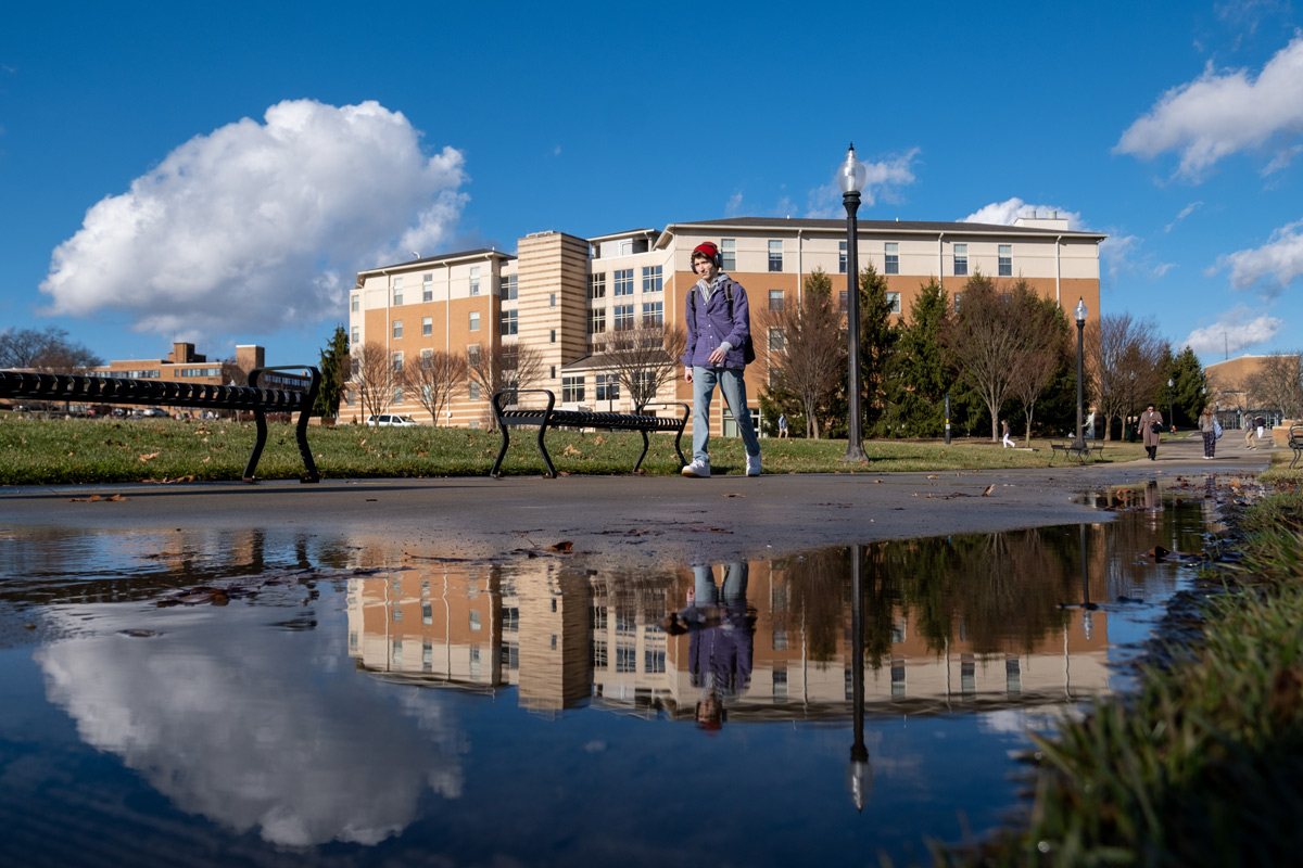 A student walking beside a puddle reflecting a residence hall and blue sky.