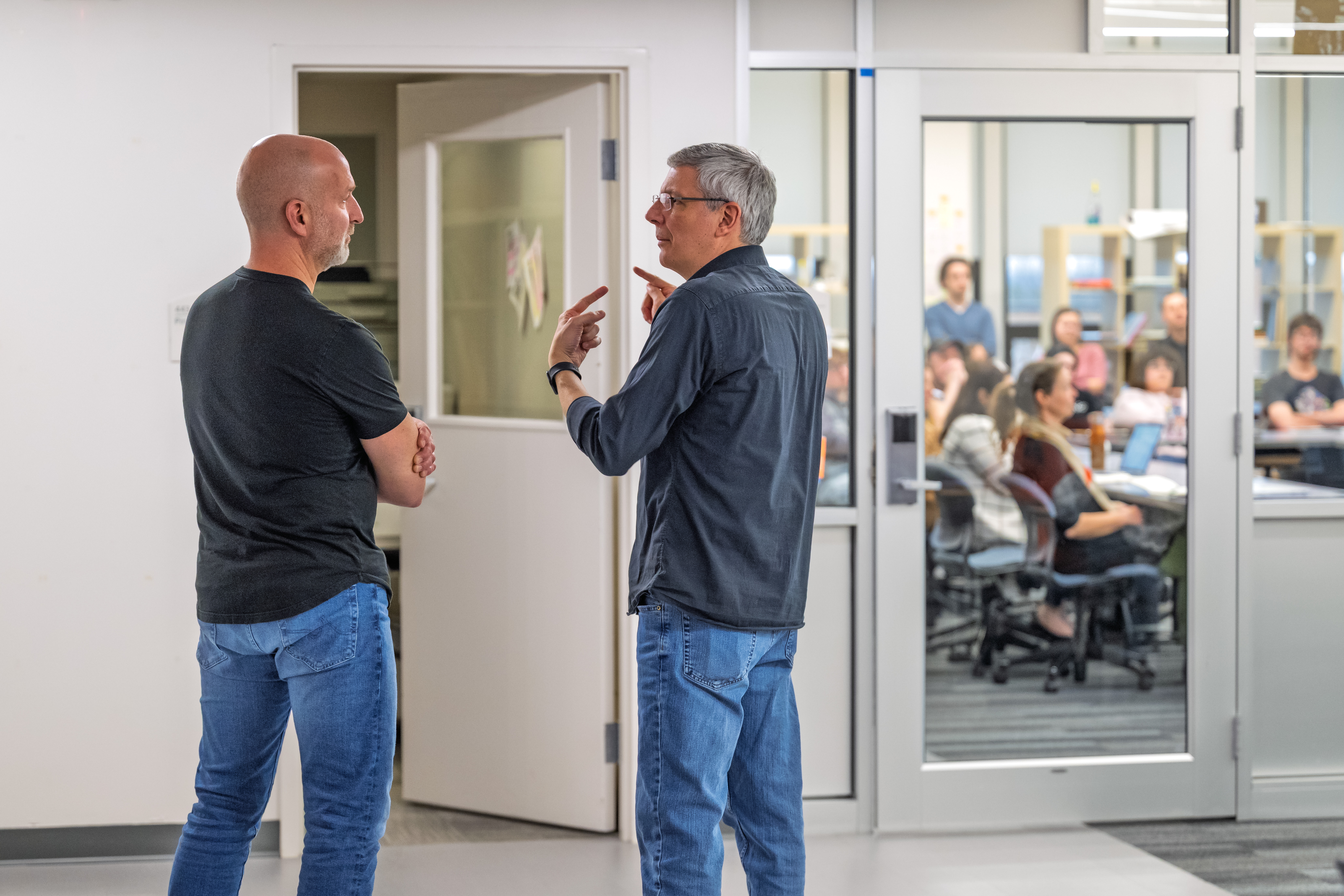 Marcus Lehto speaks with a Kent State faculty member in a hallway while a classroom full of students listens to a presentation behind glass.