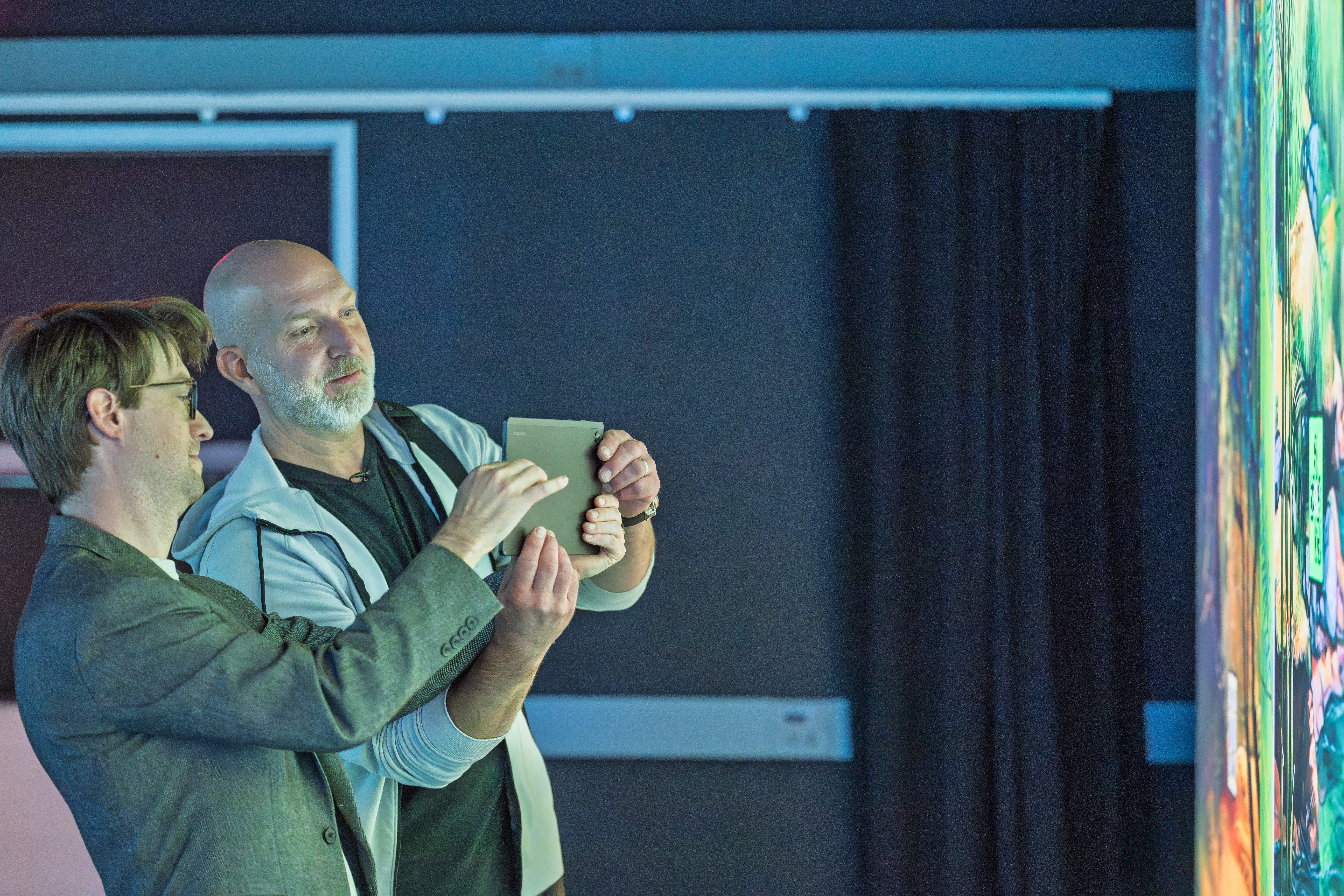 Marcus Lehto and a Kent State faculty member taking a selfie together with a tablet in a darkened room illuminated by colorful projected artwork.