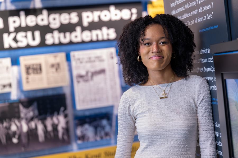 A person smiles while standing in an exhibit at the May 4 Visitors Center, with historic photos and text panels displayed behind them.