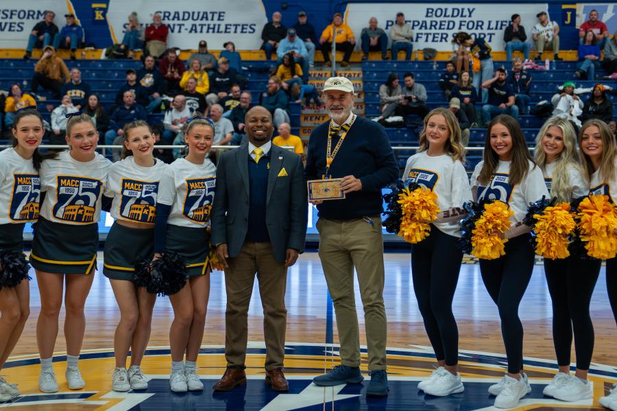 Group of cheerleaders and three adults smiling and posing at a sports event, holding an award, with spectators in the background. The group includes individuals in cheer uniforms and others in formal attire.