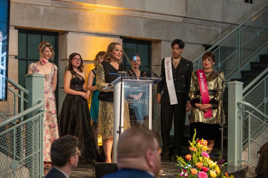 Museum director gives a speech at the dazzle gala while students stand behind her wearing best dressed sashes