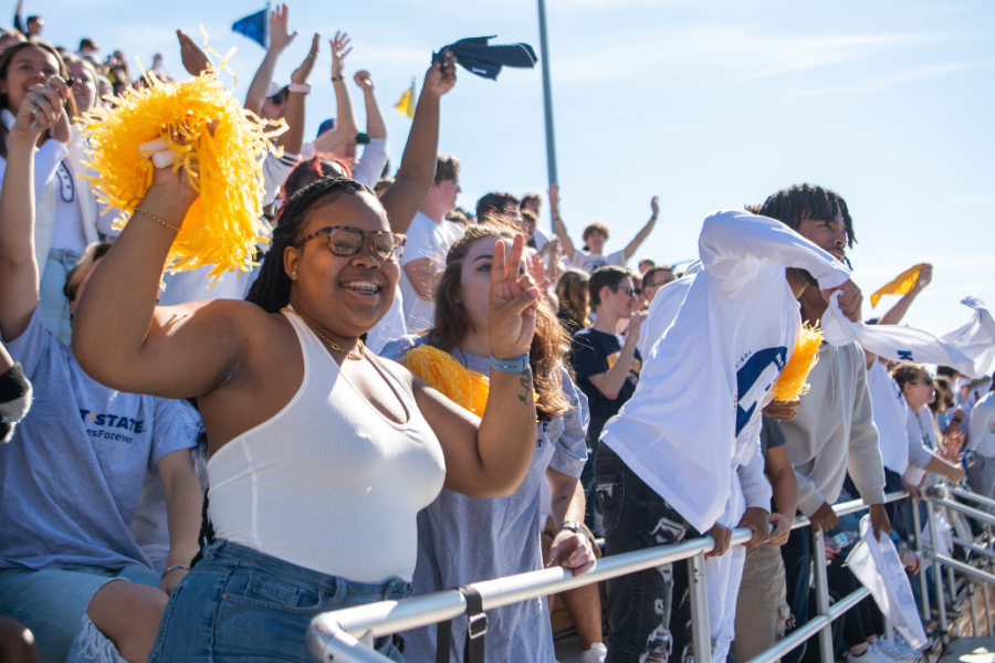 A group of student celebrate in the stands at a sporting event. One is holding a yellow pom pom.