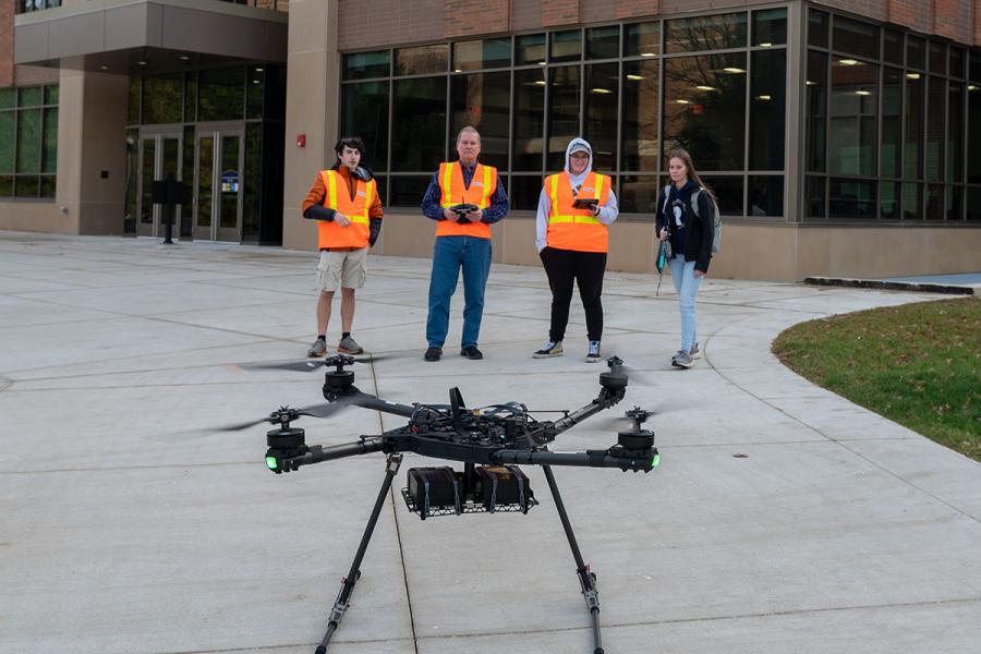 Four people standing at a distance wearing high-visibility vests with a medium-sized quadcopter in the foreground.