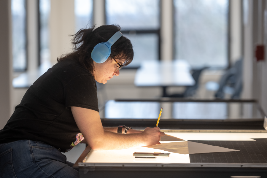 A student wearing blue headphones draws on a tracing table.