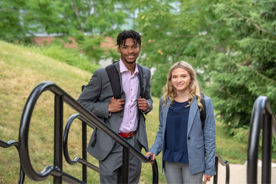 Two business students stand close together, wearing formal attire, with trees and grass in the background.
