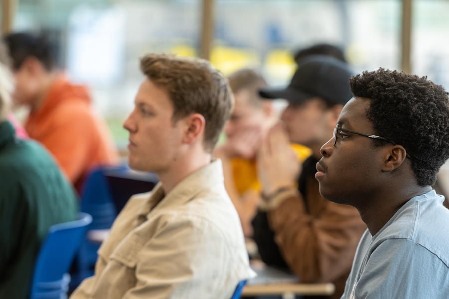A classroom of students, with a darkskinned male student in a light blue shirt sitting in front of a light skinned male student in a yellow shirt