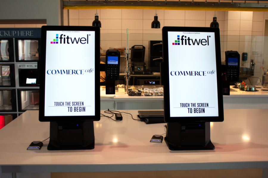 Close up of the two Commerce Cafe ordering kiosks, both featuring touchscreens, card readers, FLASHcard readers. Both screens have the Fitwel logo at the top, the Commerce Cafe logo in the center, and read "Touch the screen to begin" at the bottom.