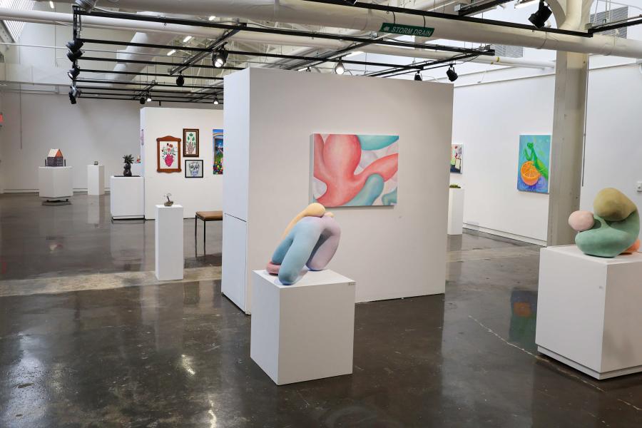 Wide view of a contemporary art gallery with polished concrete floors, white walls, and track lighting, featuring colorful abstract paintings on the walls and soft, organic-shaped sculptures displayed on white pedestals throughout the open space.