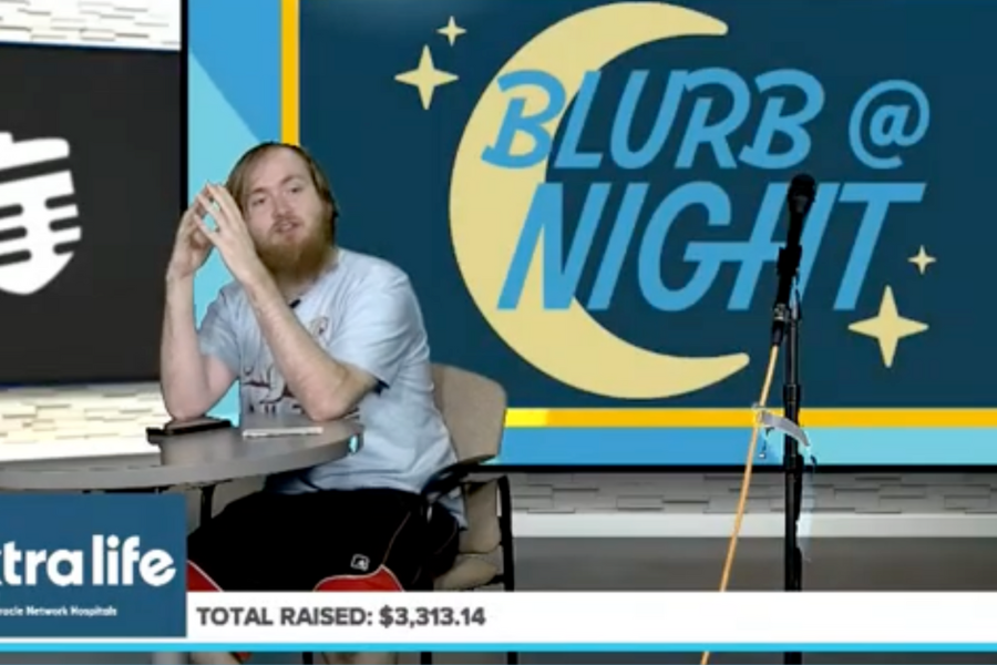 Student host sits at a studio desk during a live Extra Life broadcast, with a “Blurb @ Night” graphic behind him and a fundraising total displayed.