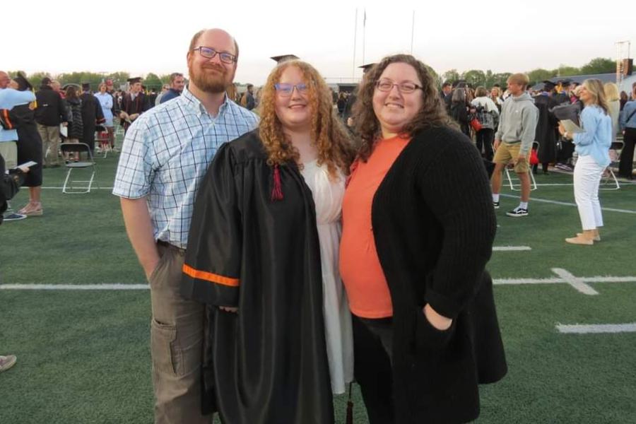 Abigail stands with her parents in her graduation gown at her high school graduation ceremony on a football field. 