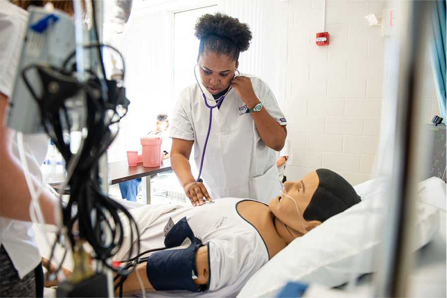A student in a lab coat talks to a woman in a hospital bed.