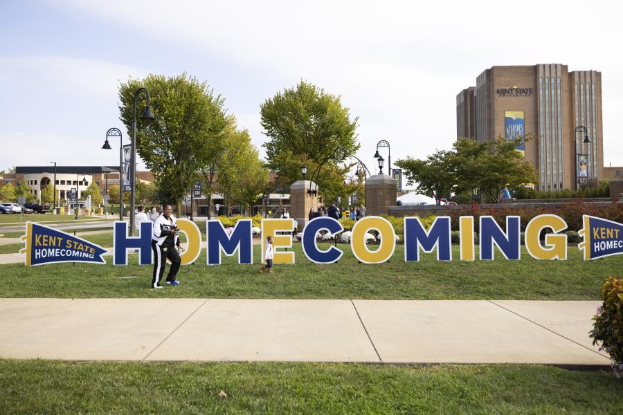 A lawn sign that reads "homecoming" in front of kent state's campus with 2 people walking in front of it