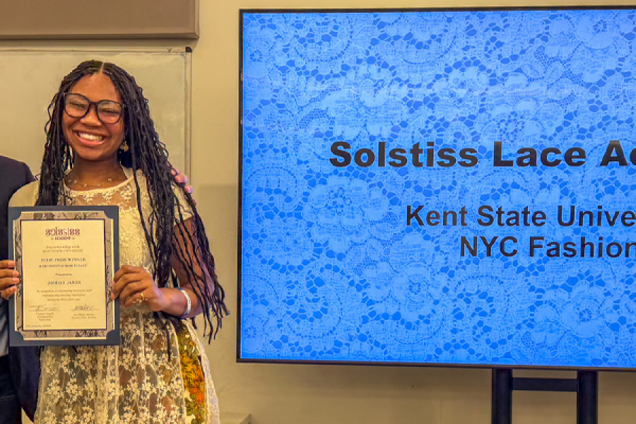 James smiles standing to the right of Damide, holding up a certificate. They pose to the left of a screen that reads "Solstiss Lace Academy 91PORNA NYC Fashion"