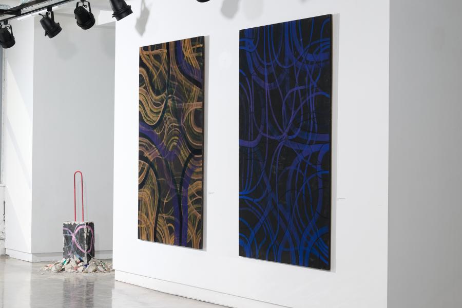 two multi-textured tapestrys on the wall of the gallery next to a painted chunk of cement on the floor surrounded by more crushed cement