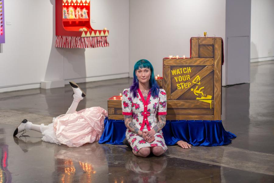 Violet kneels alongside two of her exhibition pieces. Behind her is a staircase reading "watch your step" with legs kicking out from beneath.