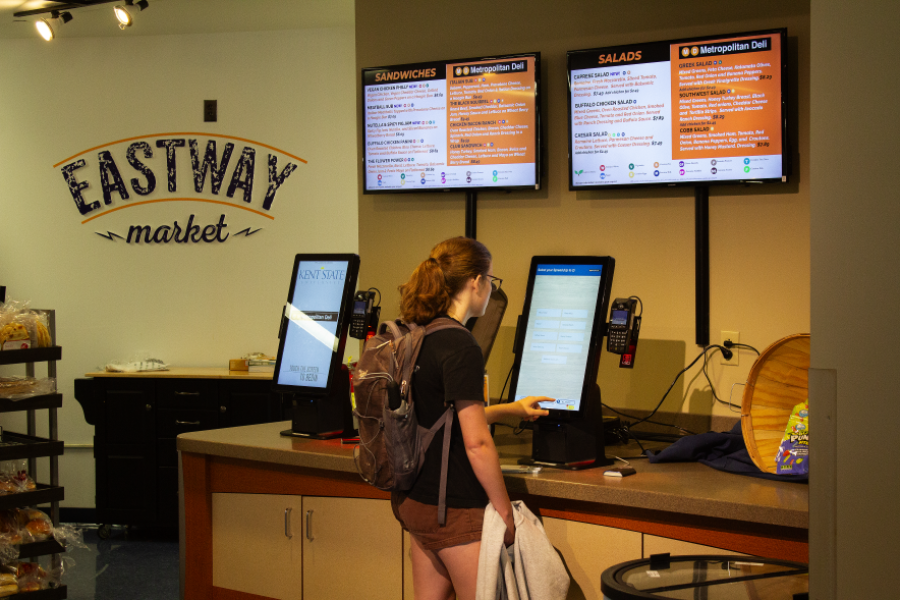 Student putting in their order at the order kiosk counter at Metro Deli, with menu screens visible above the counter