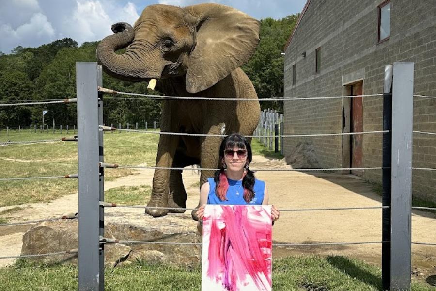 Natalie Holland standing in front of the elephant Sukiri holding a painting.